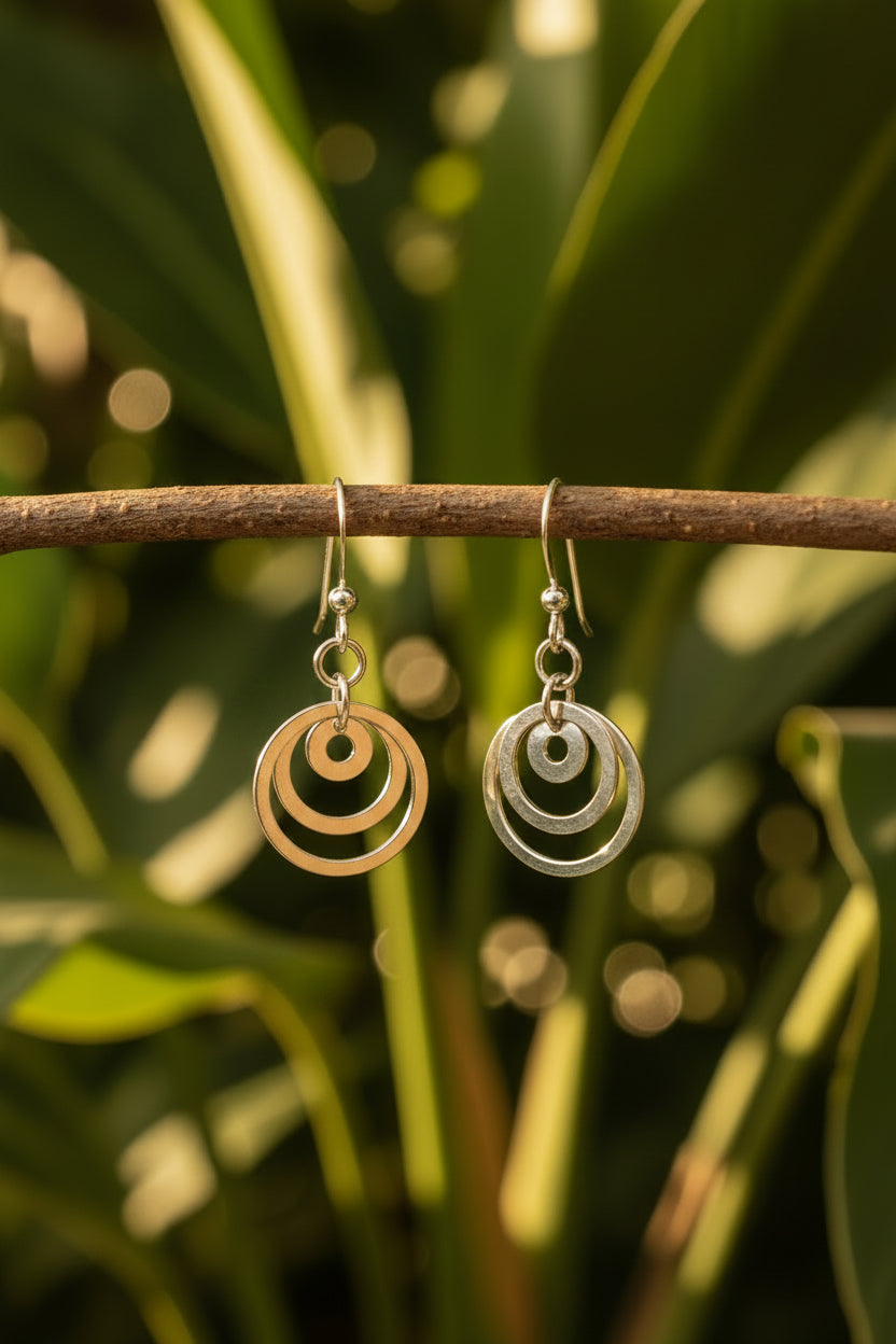 A pair of silver dangle earrings with concentric circular cutout designs, displayed on a twig with greenery plants in the background. Cheeky Goddess.