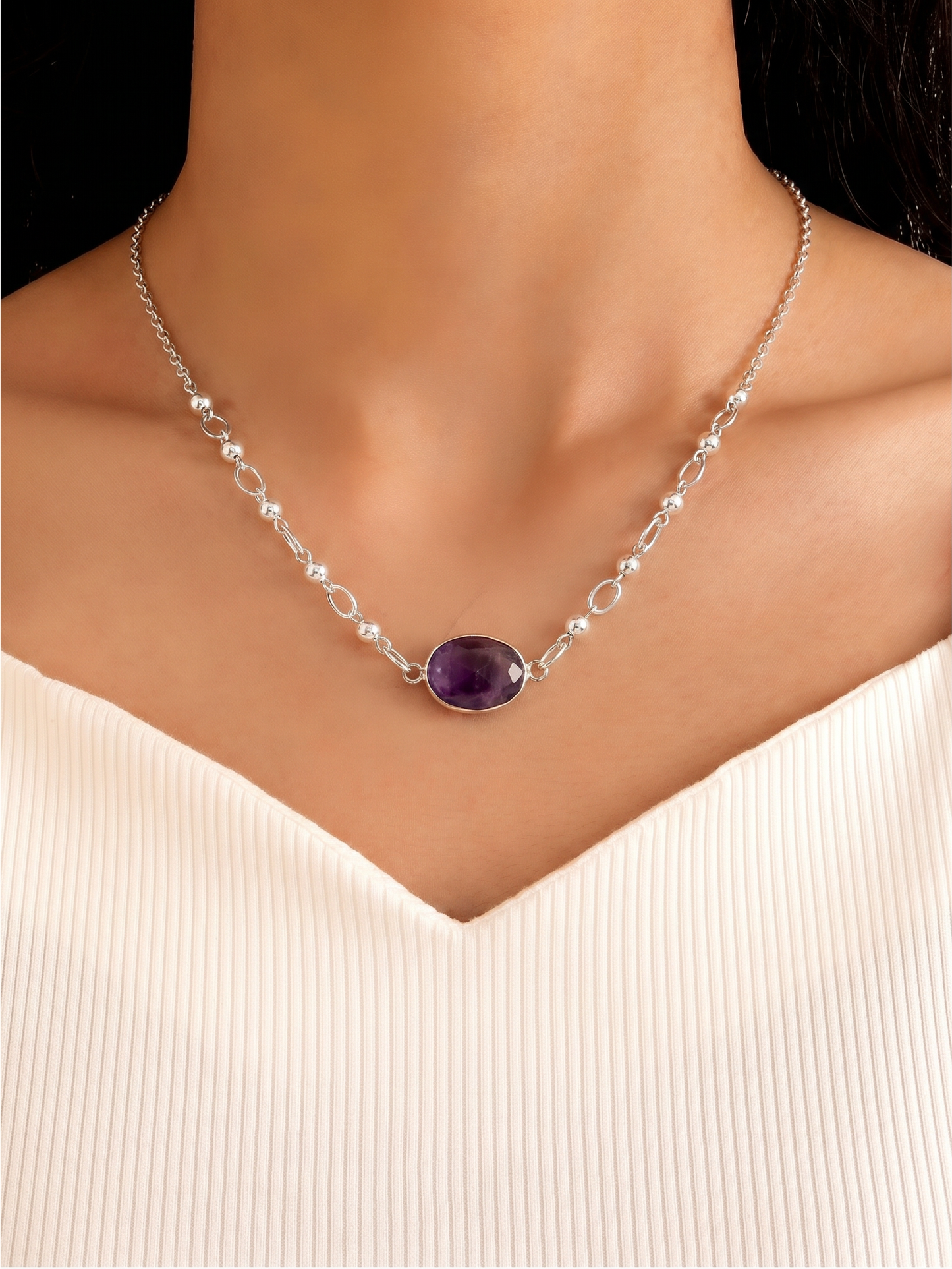 A close-up of a model's neck and upper chest wearing a delicate silver chain necklace with a central oval purple gemstone pendant.