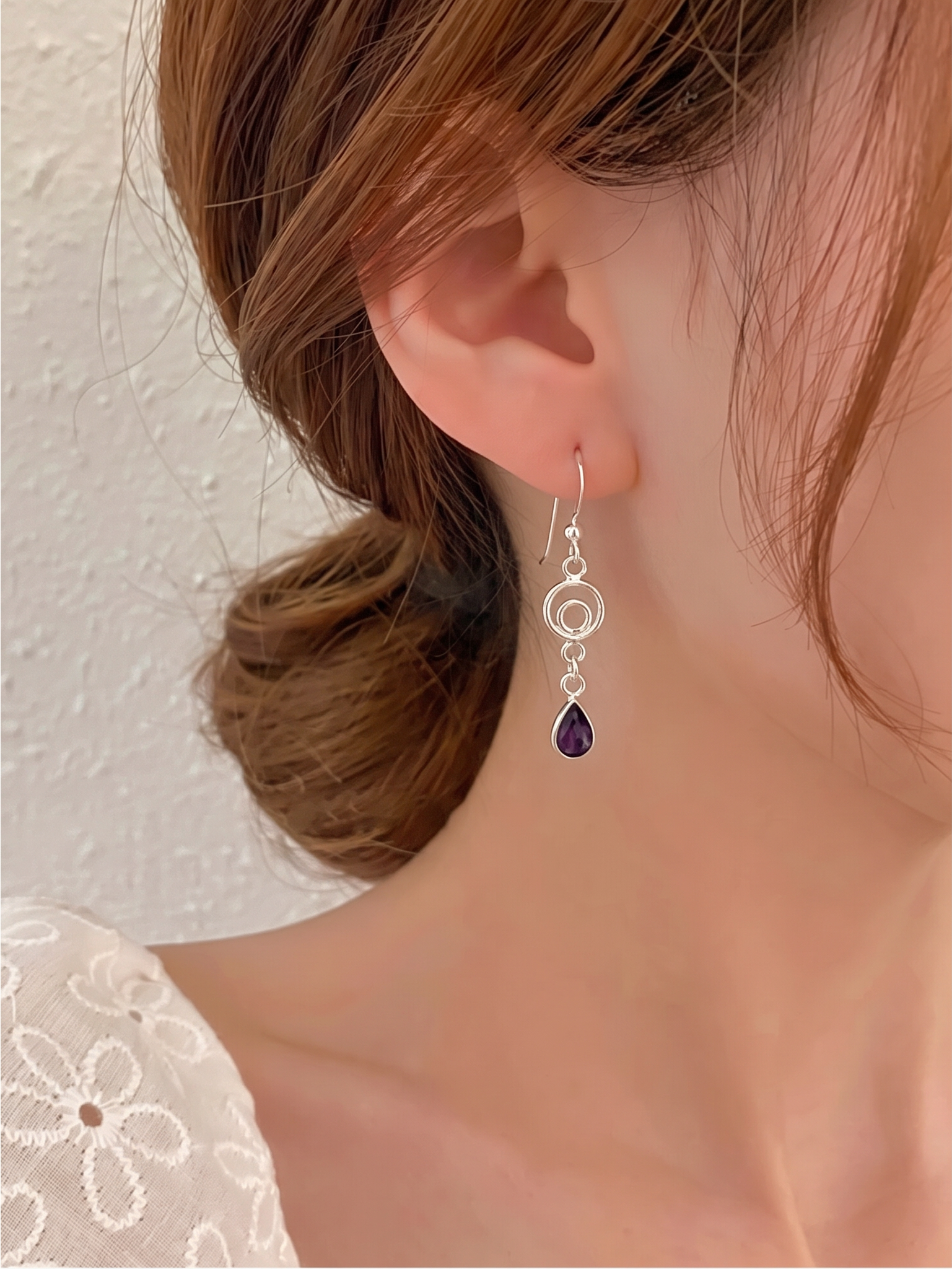 A close-up of a woman's ear wearing a silver dangle earring with a teardrop-shaped dark purple gemstone, against a textured off-white wall.