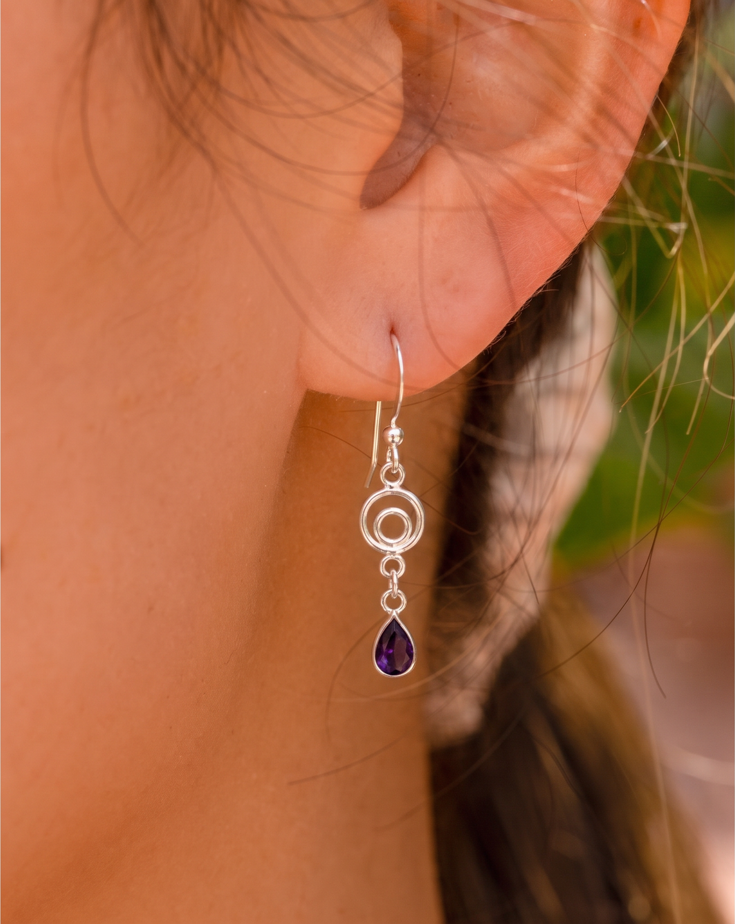 A close-up of a person's ear wearing a silver dangle earring with a teardrop-shaped purple gemstone.