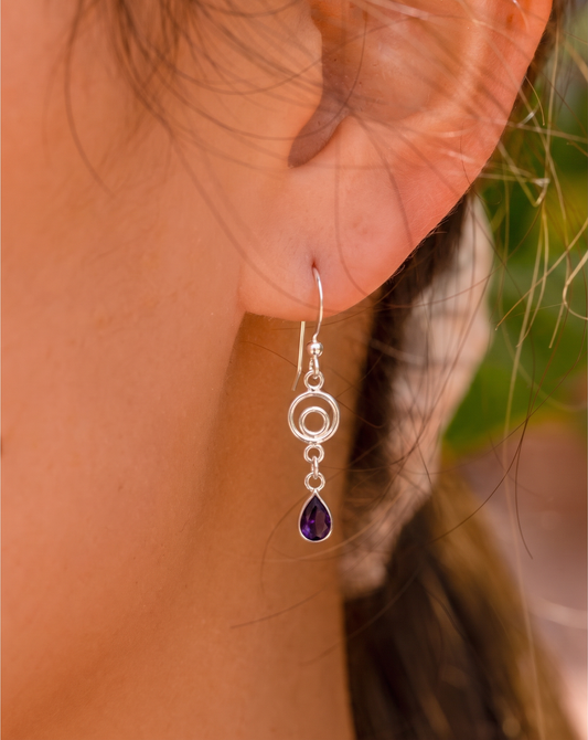 A close-up of a person's ear wearing a silver dangle earring with a teardrop-shaped purple gemstone.