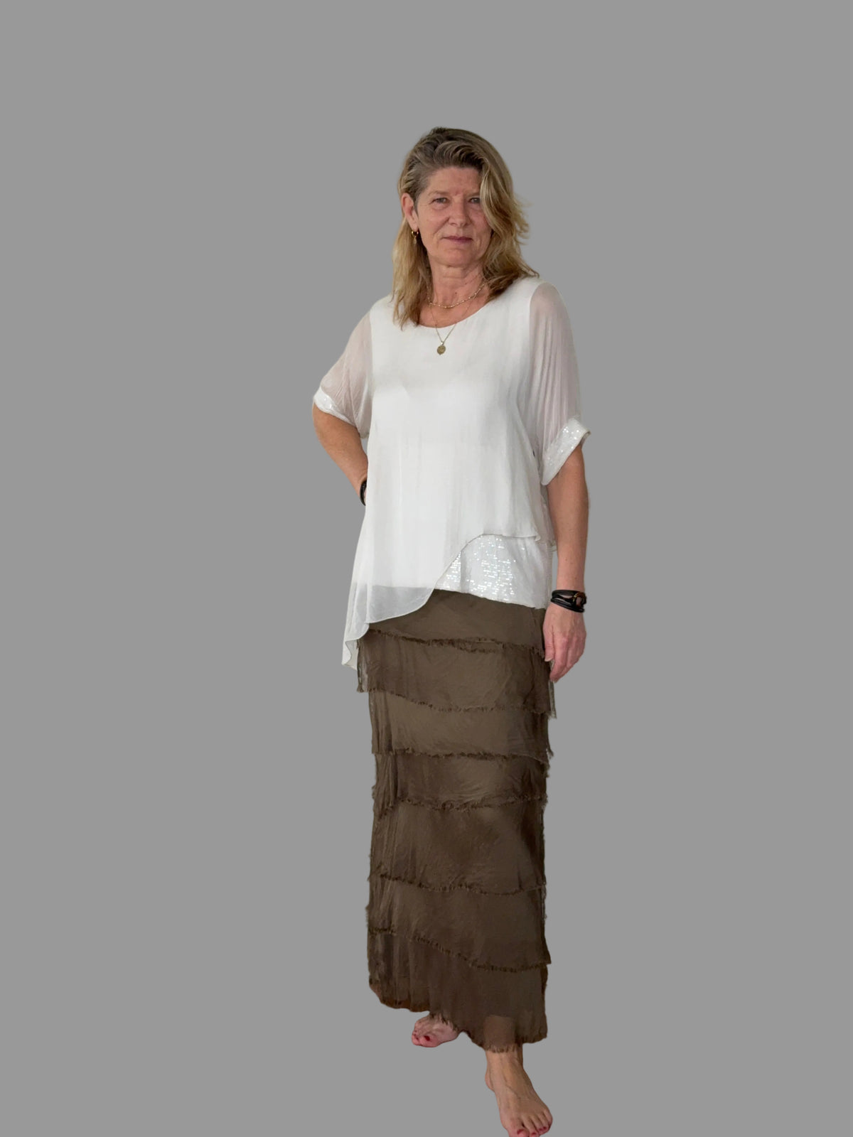 A woman modeling a white short-sleeve top with a sheer overlay and a brown tiered maxi skirt, standing against a solid gray background.