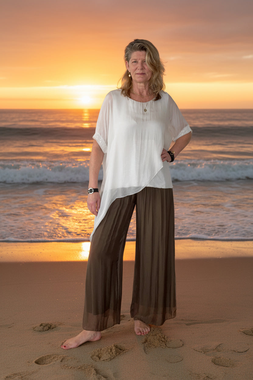 A woman stands barefoot on a wooden floor against a plain off-white wall, wearing a loose white short-sleeve top and dark brown wide-leg pleated pants.