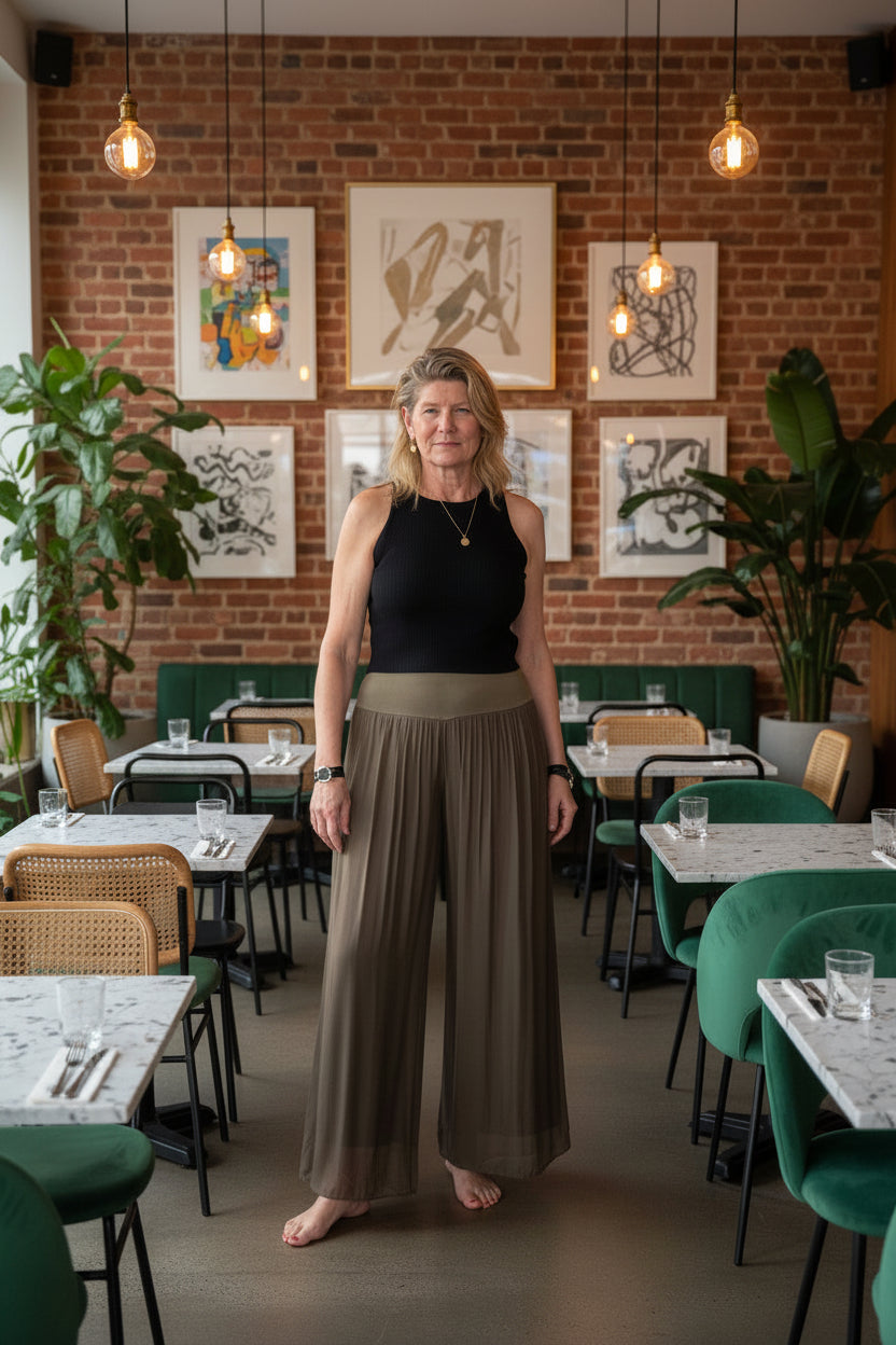 A woman stands barefoot on a wooden floor against a plain white wall, wearing a sleeveless black top and high-waisted, wide-leg taupe-brown pleated pants.