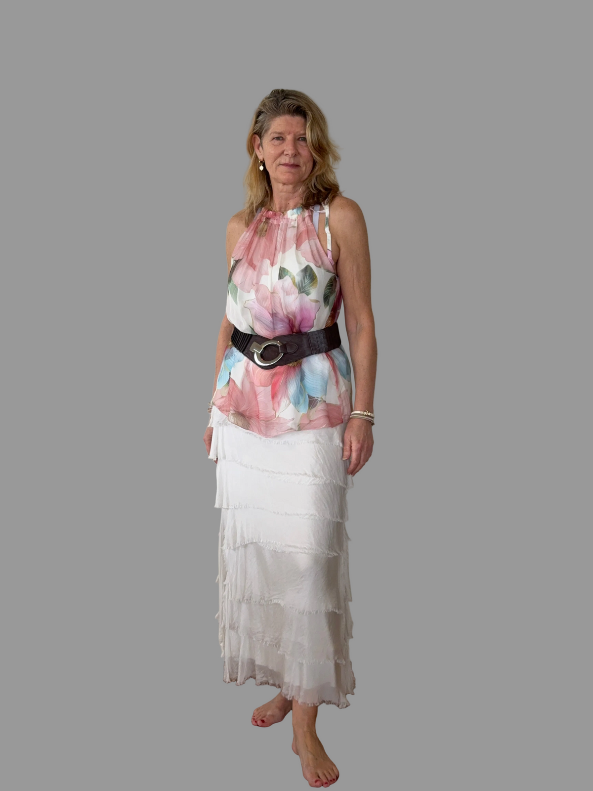 A woman modeling a sleeveless halter-neck top with a pink, blue, and green floral print, paired with a wide black belt and a long white tiered skirt, set against a solid gray background.