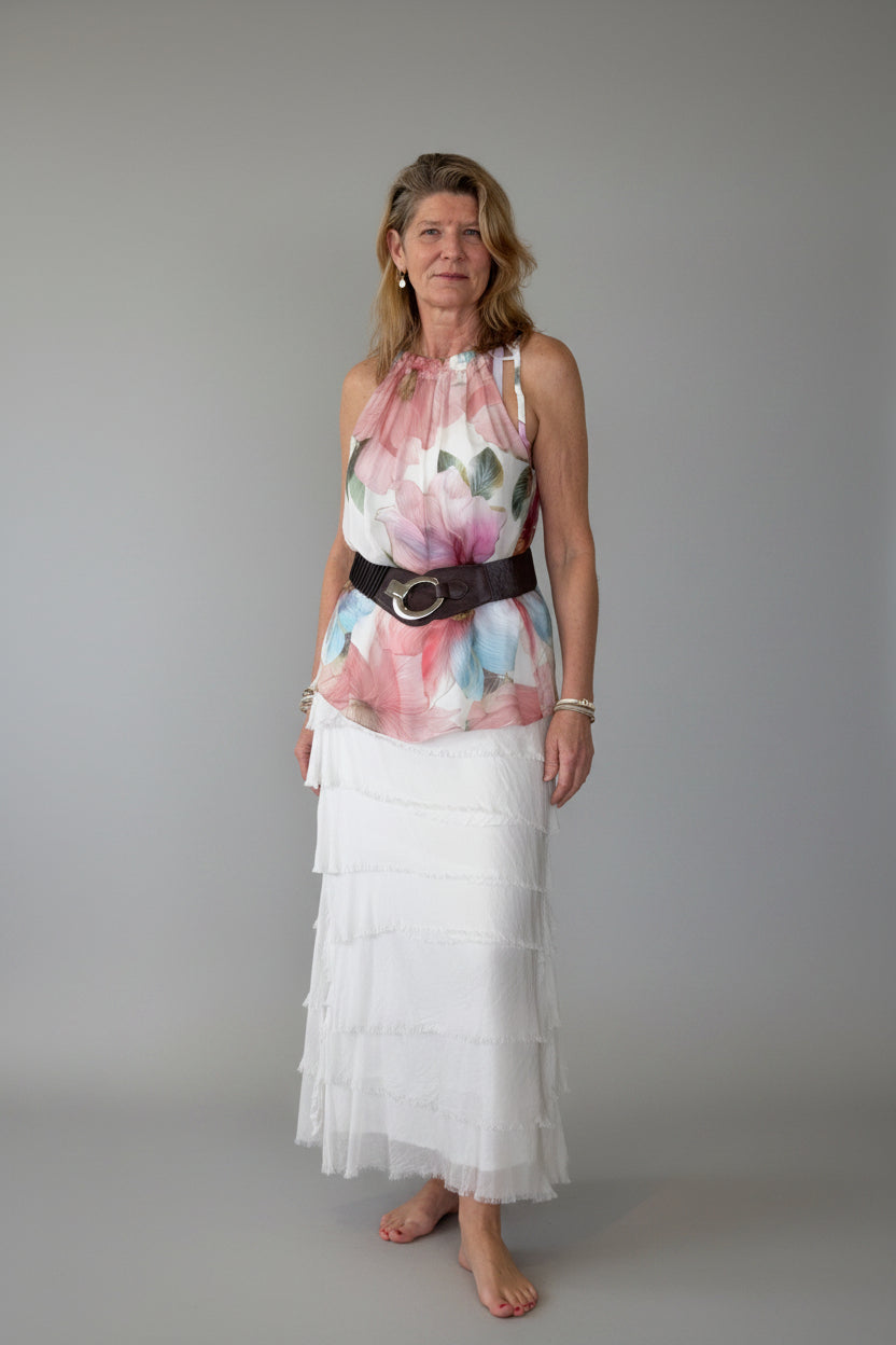 A woman stands barefoot in a room with a light grey wall, wearing a sleeveless silk long top with a pink, blue, and white floral print, a wide brown belt with a silver buckle, and a long white tiered silk skirt.  Cheeky Goddess.  Australia