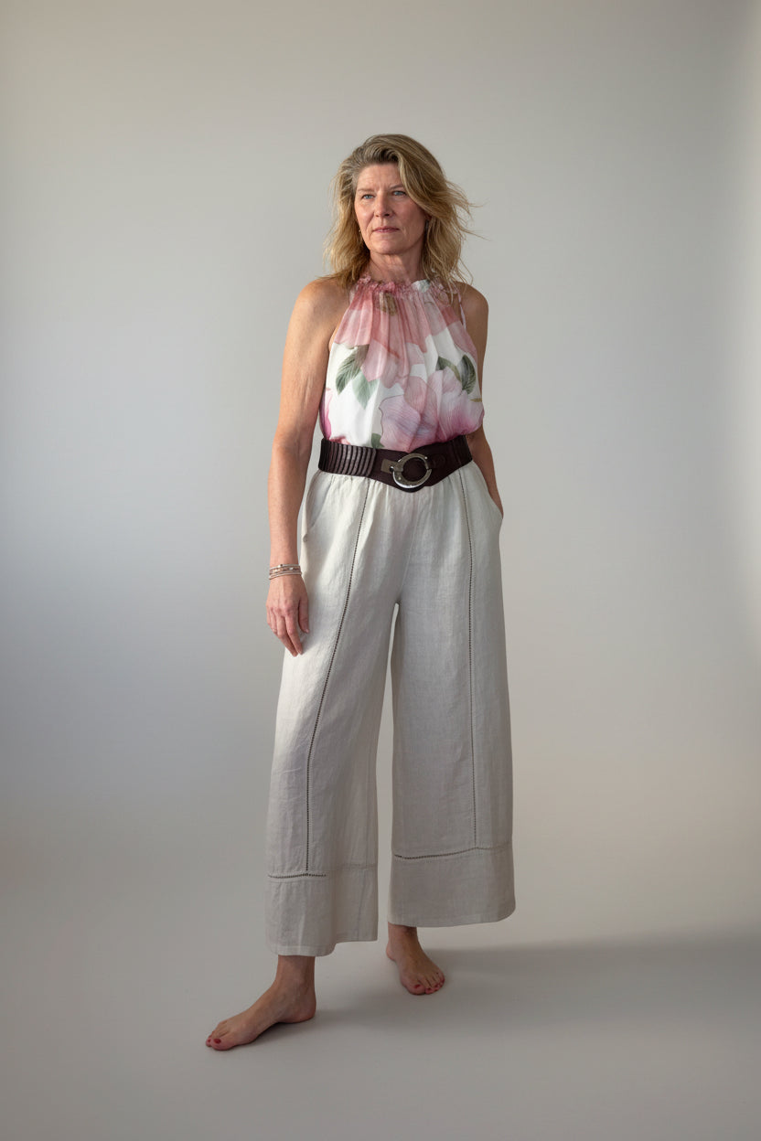 A woman stands barefoot on against a plain grey wall, wearing a sleeveless floral silk top with pink and green and cream colors tucked into linen pants with a brown belt and light beige wide-leg cropped pants.  Cheeky Goddess. Australia.