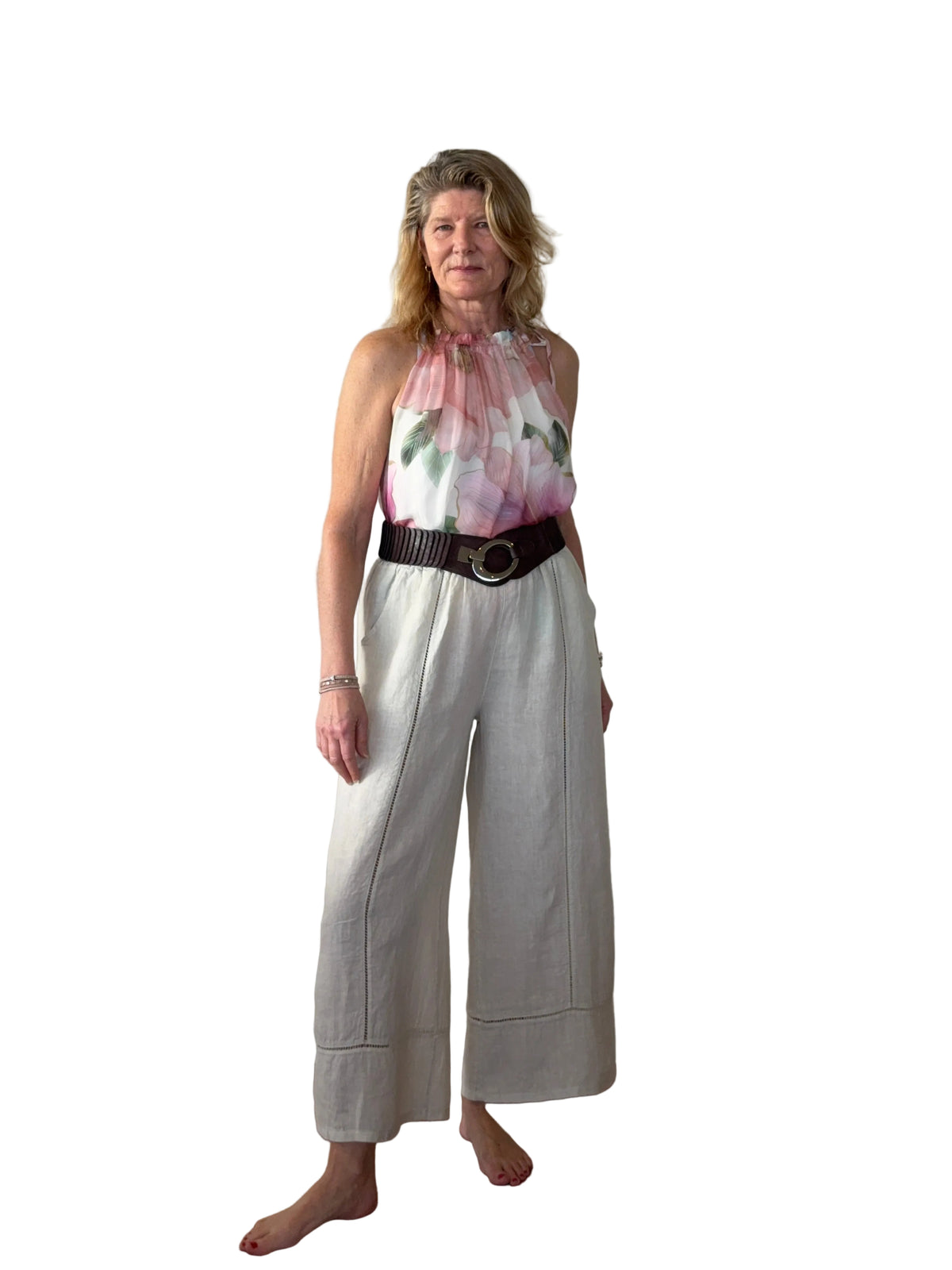 A woman stands barefoot on a wooden floor against a plain white wall, wearing a sleeveless floral halter top with a black belt and light beige wide-leg cropped pants.
