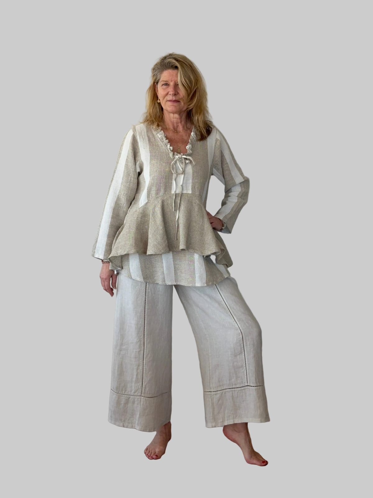 A woman modeling a two-piece linen outfit consisting of a long-sleeve peplum top with a tie neckline and matching wide-leg cropped pants, standing on a wooden floor against a plain off-white wall.