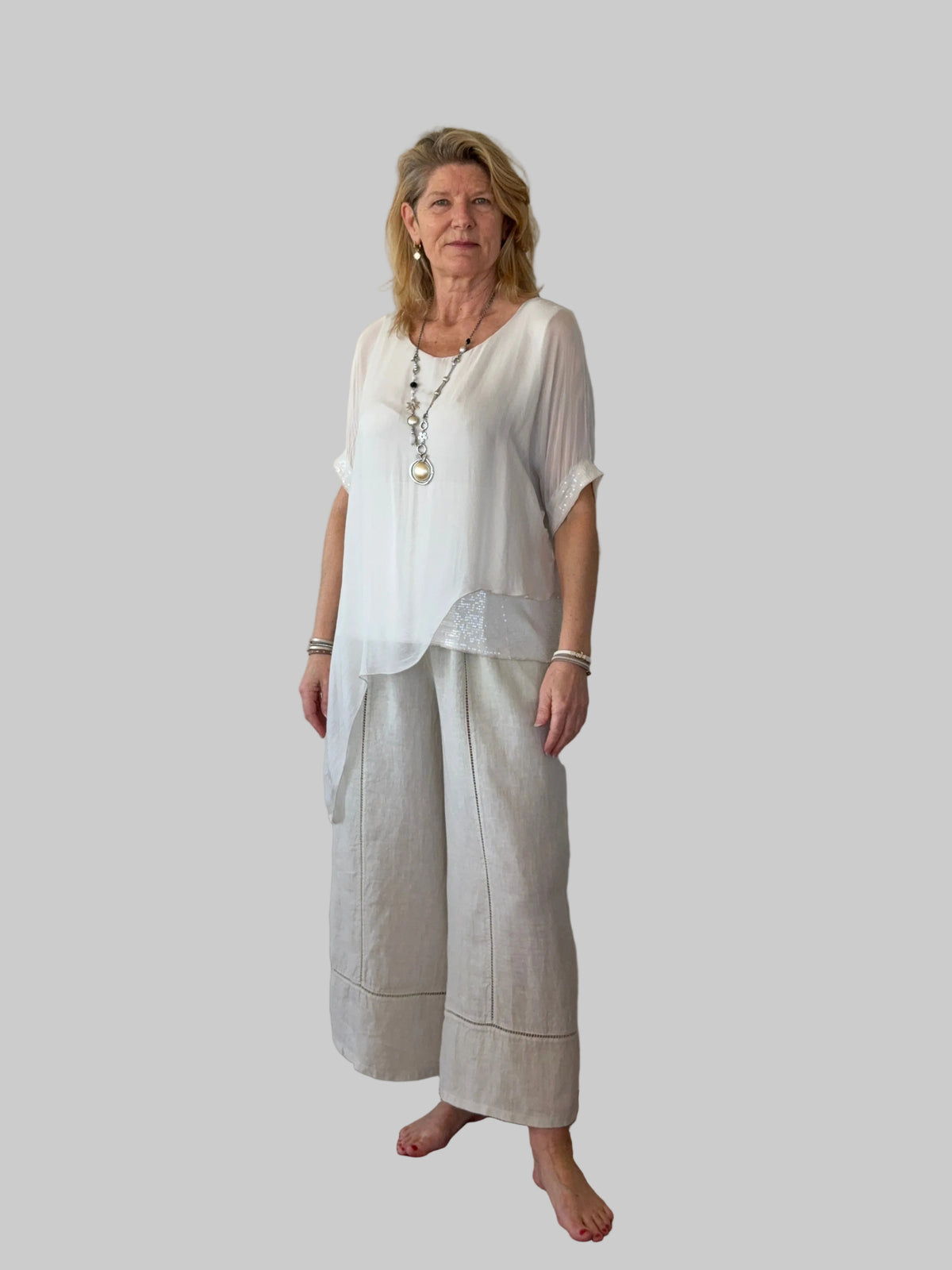 A woman stands barefoot against a plain off-white wall on a wooden floor, wearing a loose white short-sleeve top and light beige wide-leg linen pants. cheeky goddess