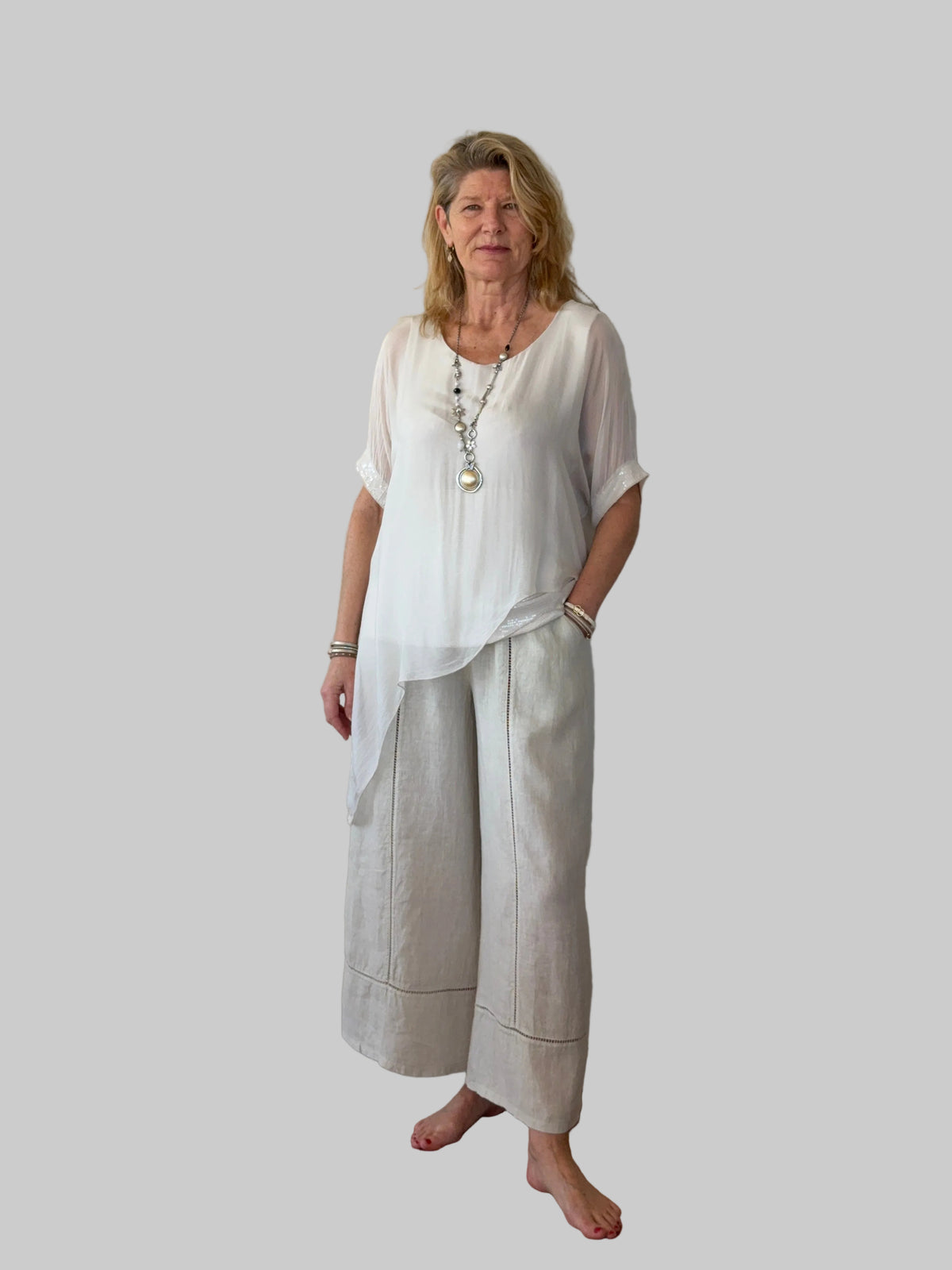 A woman stands barefoot against a plain off-white wall on a wooden floor, wearing a loose white short-sleeve top and light beige wide-leg linen pants. cheeky Goddess
