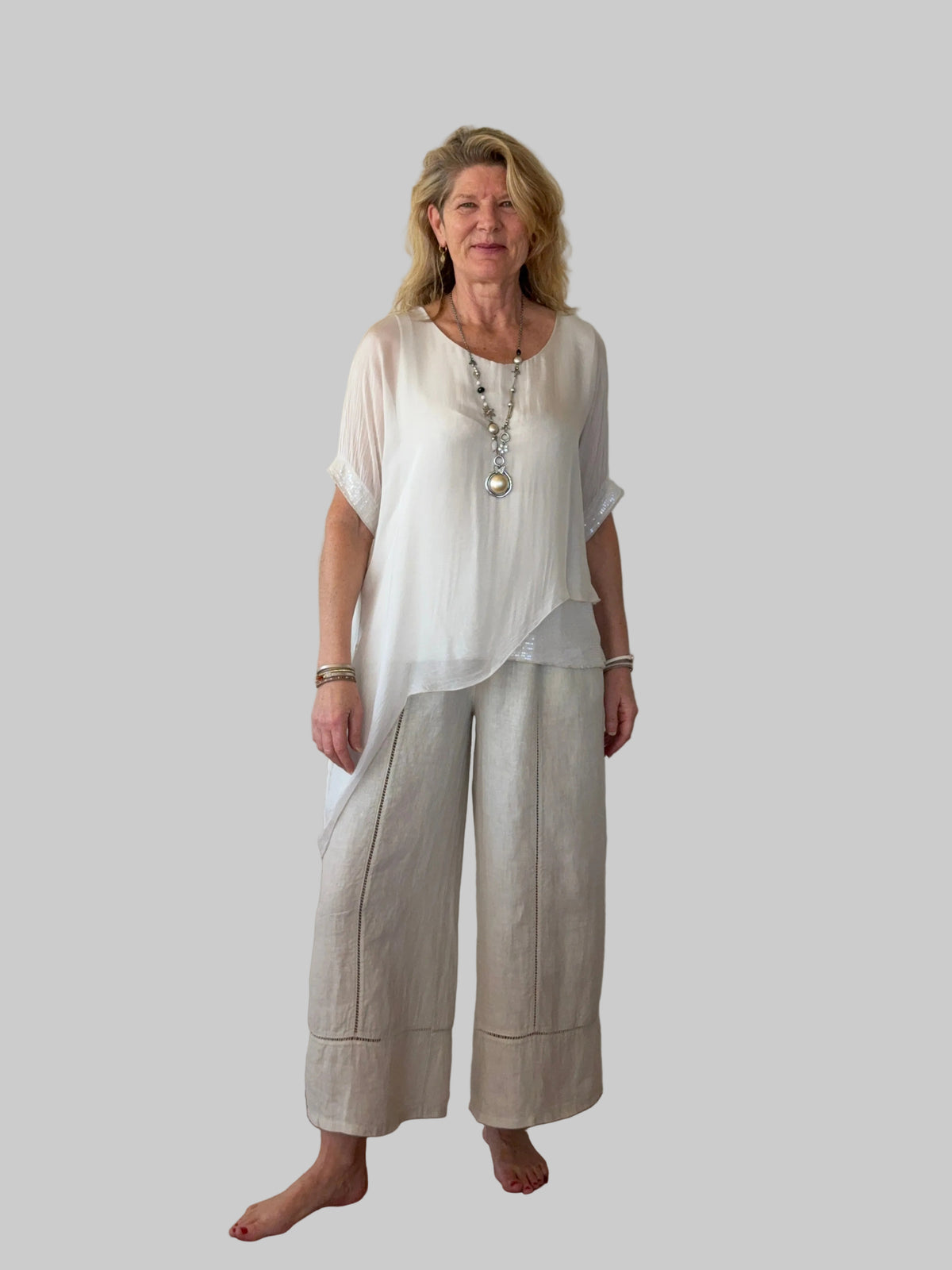 A woman stands barefoot on a wooden floor against a plain off-white wall, wearing a loose white short-sleeve top and light beige linen wide-leg pants.  Cheeky Goddess.