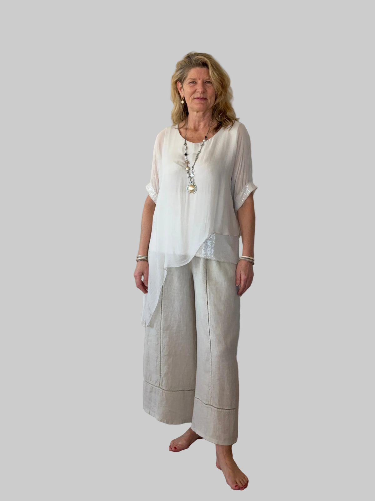A woman stands barefoot against a plain off-white wall on a wooden floor, wearing a loose white short-sleeve top and light beige wide-leg pants with a matching draped overlay.  Cheeky Goddess.