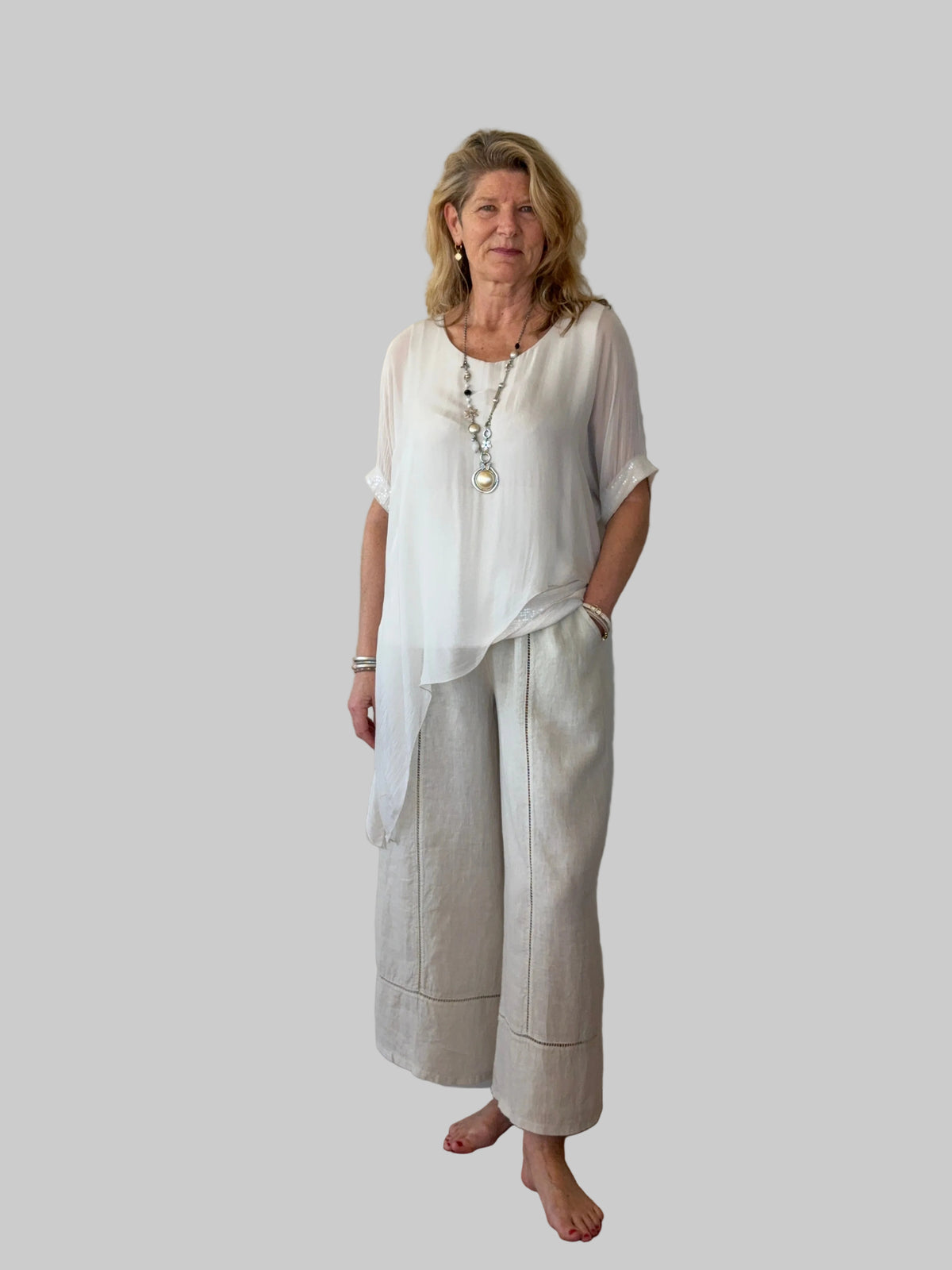 A woman stands barefoot against a plain off-white wall on a wooden floor, wearing a loose white short-sleeve top and light beige wide-leg linen pants.  Cheeky Goddess.