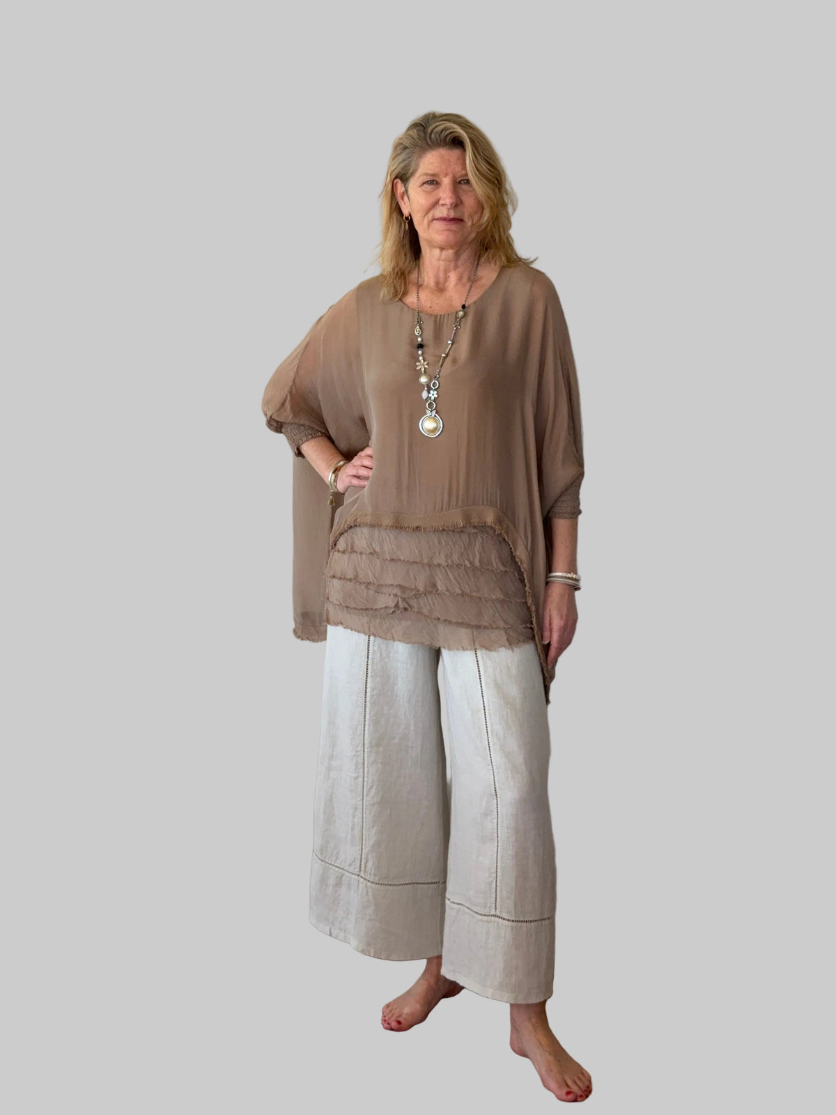 A woman stands barefoot on a wooden floor against a plain off-white wall, wearing a loose taupe-brown draped top with a ruffled hem and light beige wide-leg pants.  Cheeky Goddess.