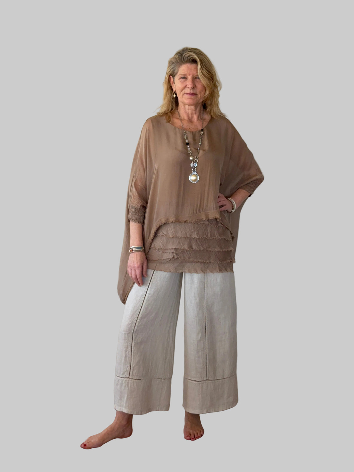 A woman stands barefoot against a plain off-white wall, wearing a loose taupe-brown sheer top with a ruffled hem and light beige wide-leg cropped pants.  Cheeky Goddess.