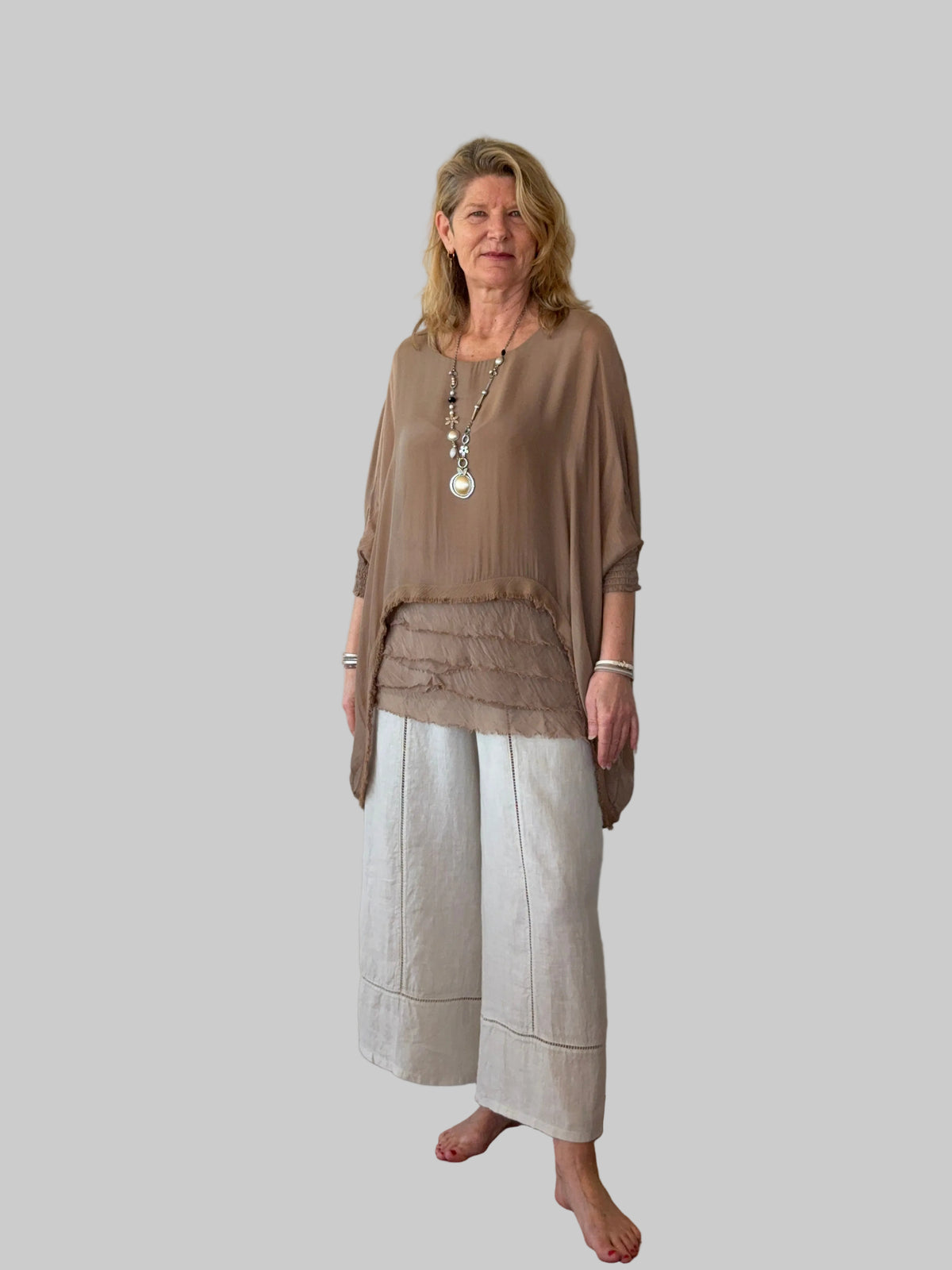 A woman stands barefoot against a plain off-white wall on a wooden floor, wearing a loose taupe-brown draped top and light beige wide-leg pants.  Cheeky Goddess.