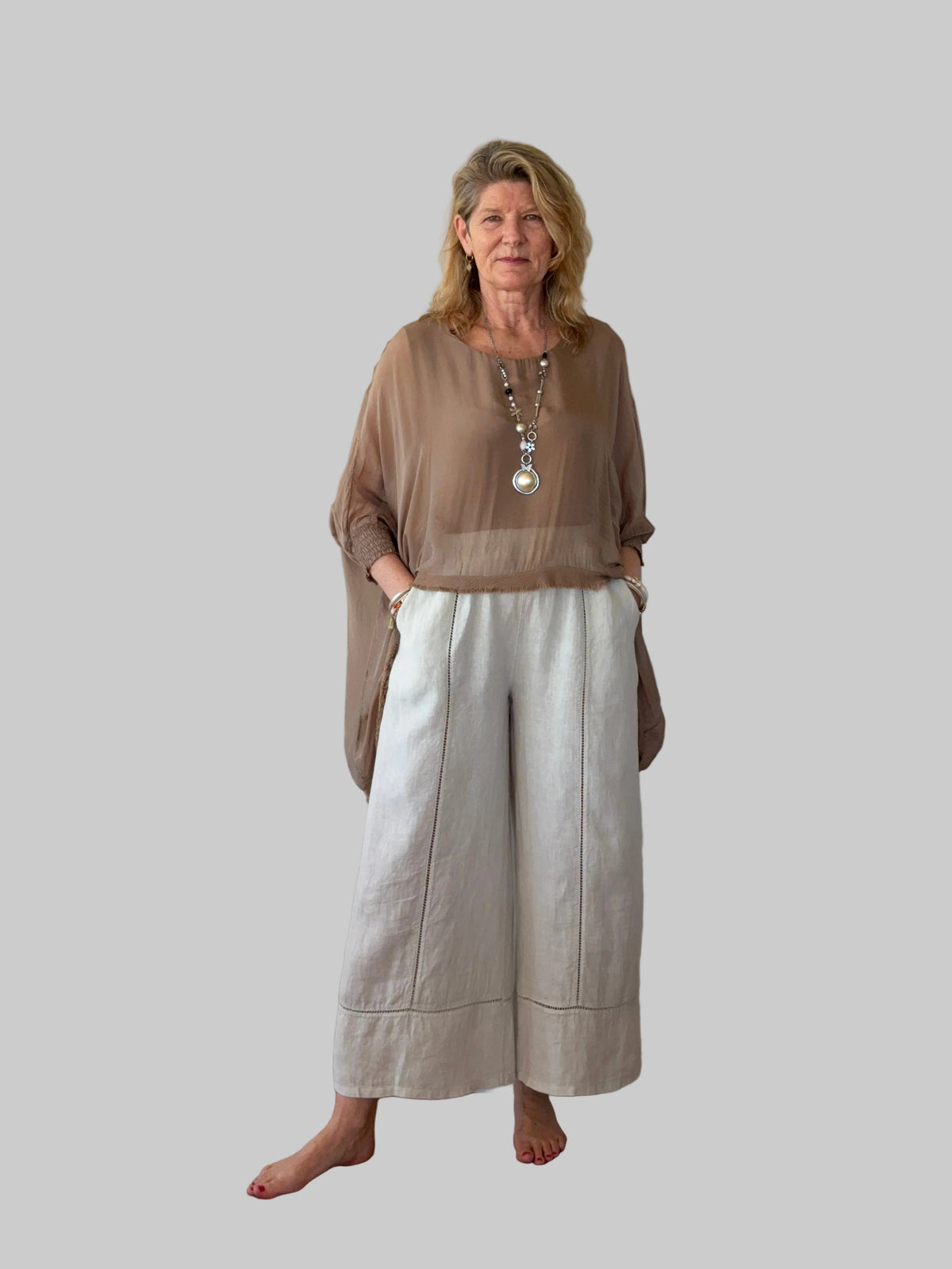 A woman stands barefoot against a plain off-white wall on a wooden floor, wearing a sheer taupe-brown long-sleeve top and light beige wide-leg linen pants.  Cheeky Goddess.