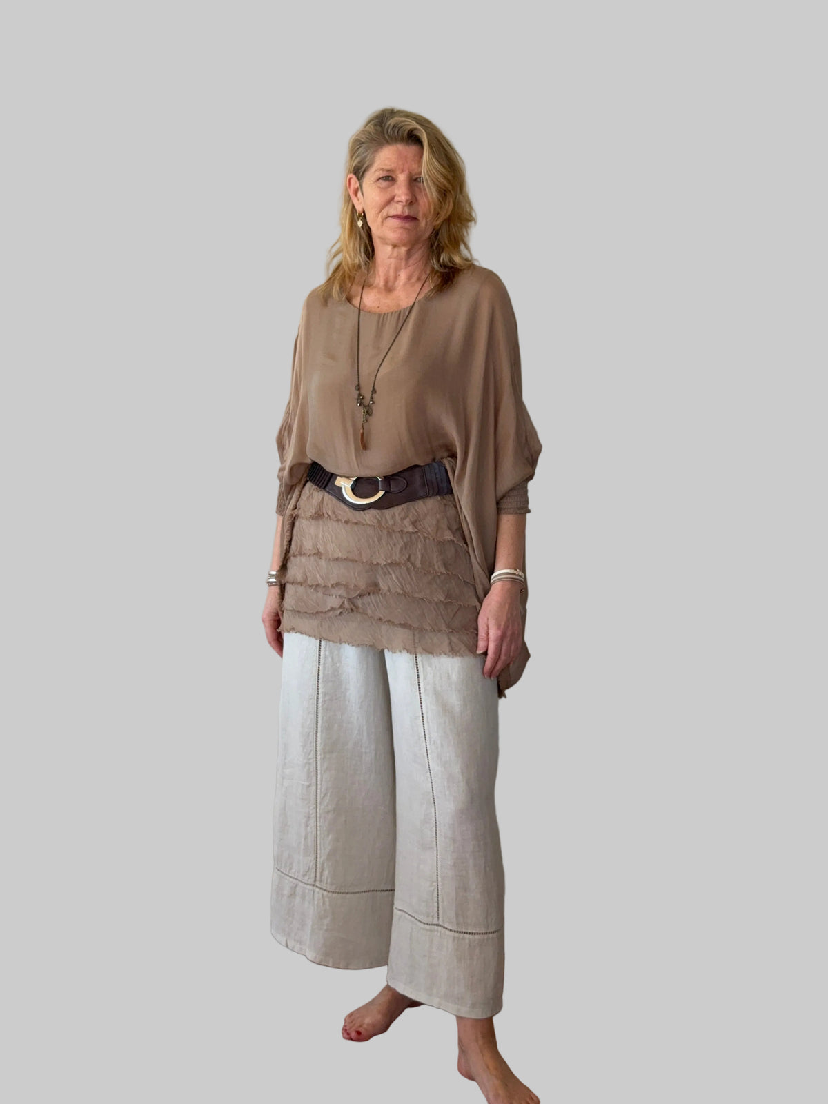 An older woman stands barefoot on a wooden floor against a plain off-white wall, wearing a loose taupe-brown top, a dark brown belt with a gold-tone ring, and light beige wide-leg cropped pants.  Cheeky Goddess.