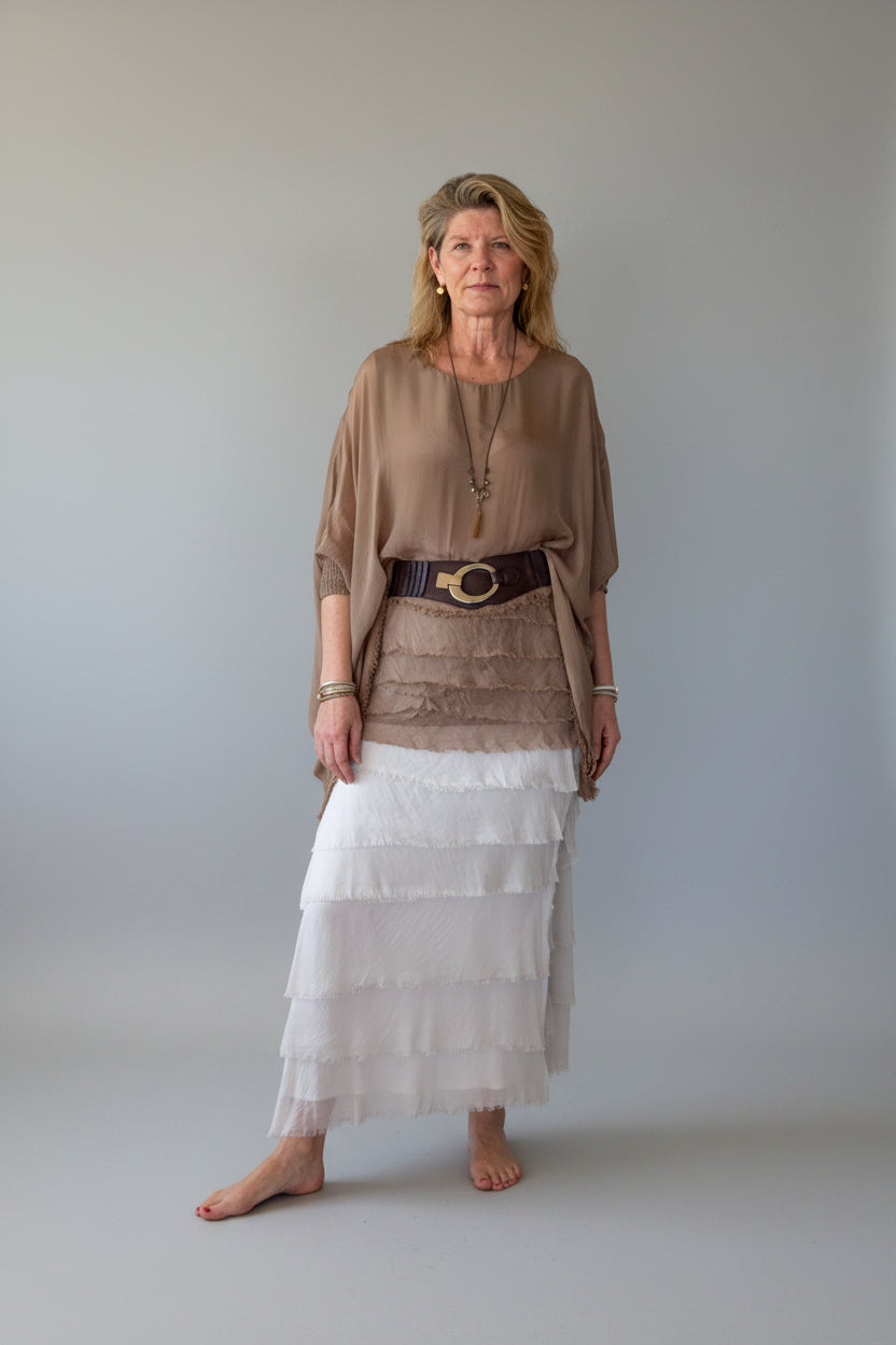 A woman stands barefoot against a plain off-white wall, wearing a loose taupe-brown silk top, a dark brown wide belt with a gold-tone buckle, and a long white tiered silk skirt.  Cheeky Goddess Australia