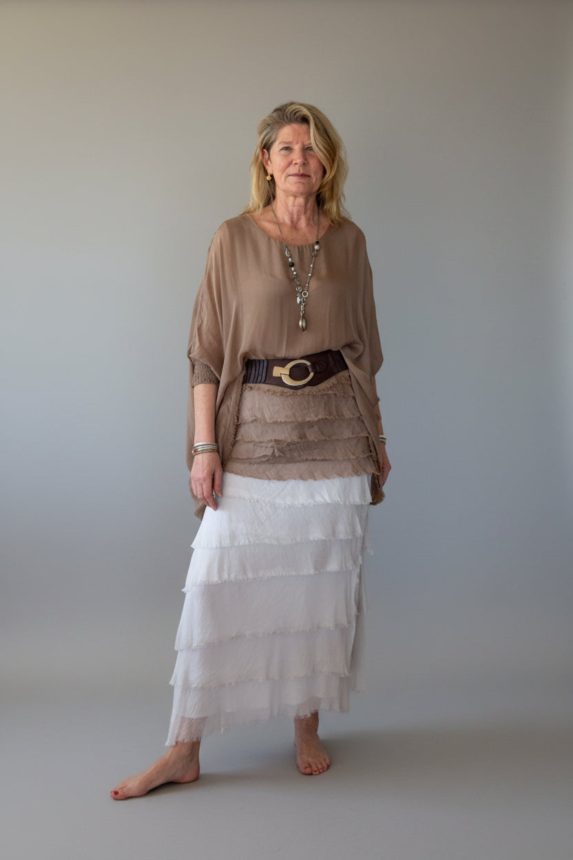 A woman stands barefoot against a plain off-white wall, wearing a loose taupe-brown silk top, a dark brown belt with a gold buckle, and a long white tiered silk skirt.  Cheeky Goddess, Australia.