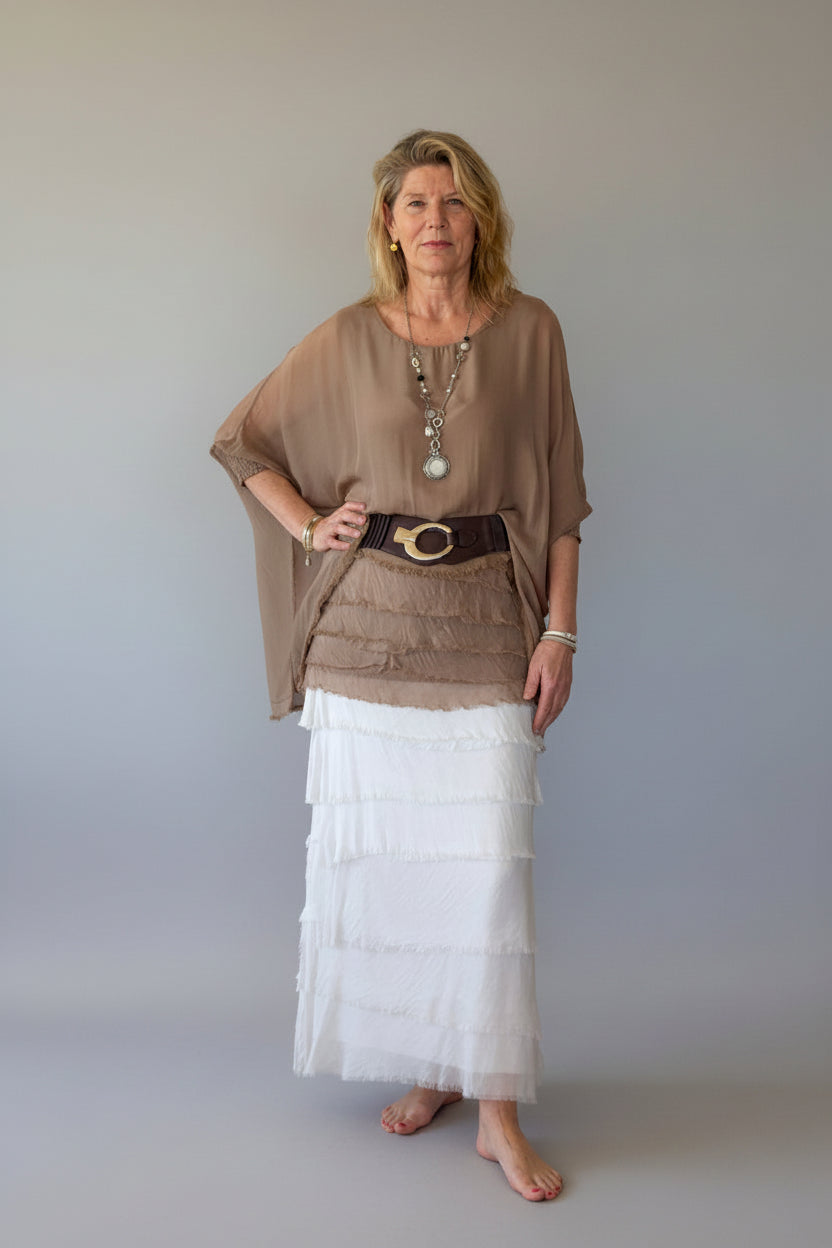 A woman stands barefoot against a plain grey wall, wearing a loose taupe-brown tunic silk top with a wide dark belt, and a long white tiered silk skirt.  Cheeky Goddess.  Australia