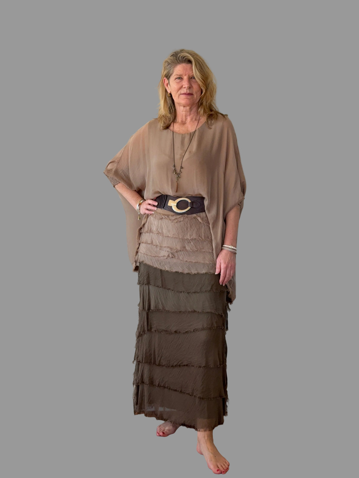 A woman stands barefoot against a plain off-white wall, wearing a loose taupe-brown short-sleeve top, a wide dark belt with a gold buckle, and a long tiered skirt in taupe and brown.