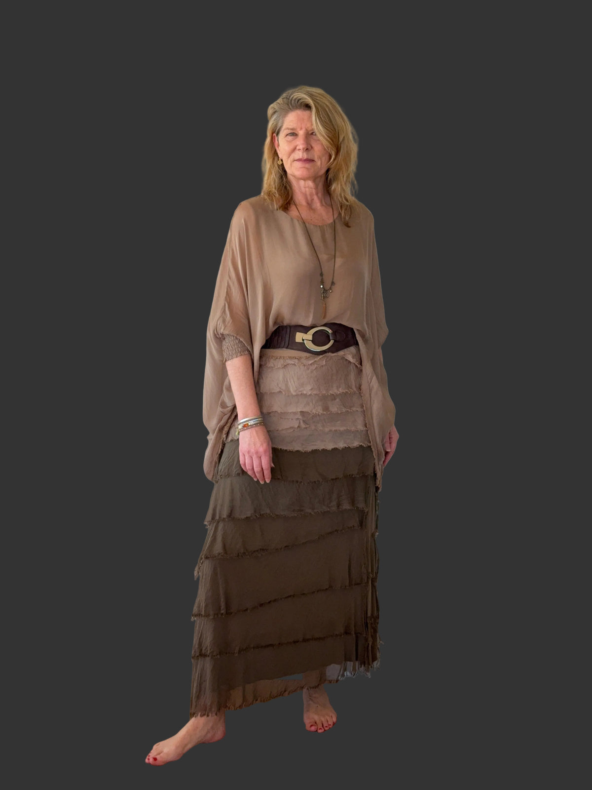 An older woman stands barefoot on a wooden floor against a plain off-white wall, wearing a layered bohemian-style outfit consisting of a taupe top, a dark brown belt, a sheer beige waist sash, and a long dark brown ruffled tiered skirt.