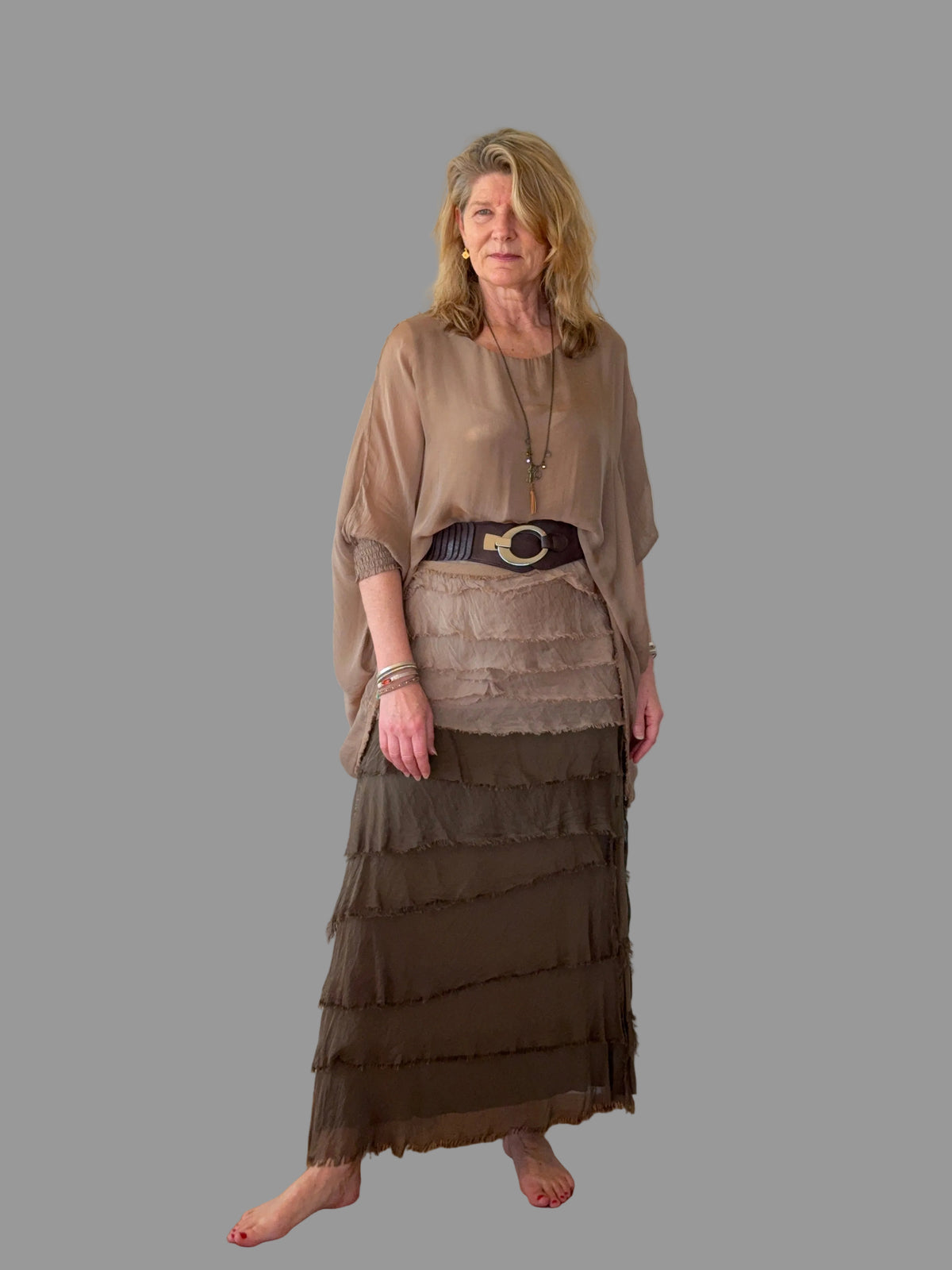 An older woman stands barefoot on a wooden floor against a plain off-white wall, wearing a layered taupe and brown dress with a wide dark belt featuring a large gold buckle.