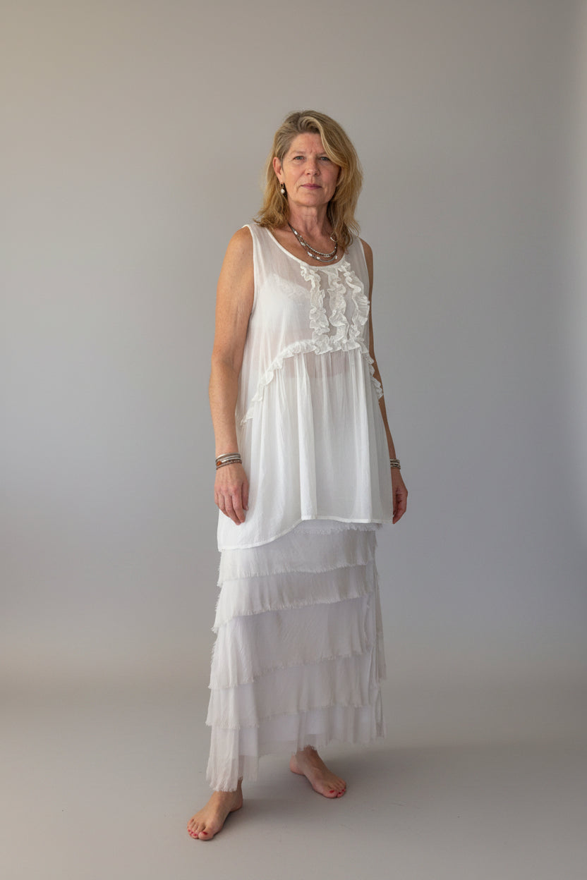 A woman modeling a white sleeveless long cotton top with a ruffled neckline and a latte colored silk tiered skirt, standing against a plain off-white wall.  Cheeky Goddess.  Australia