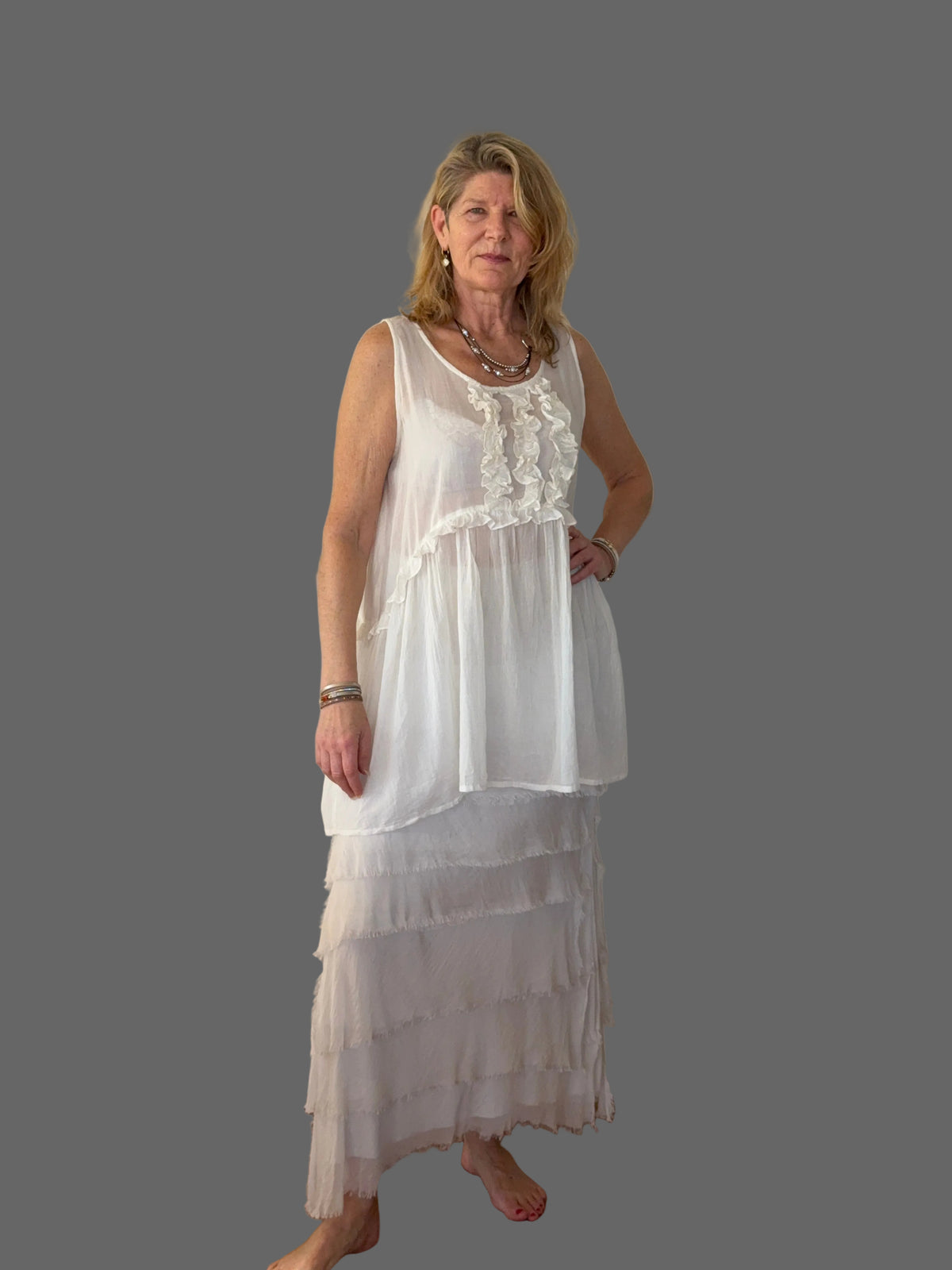 A woman modeling a white sleeveless maxi dress with a ruffled neckline and a tiered skirt, standing against a plain off-white wall on a wooden floor.