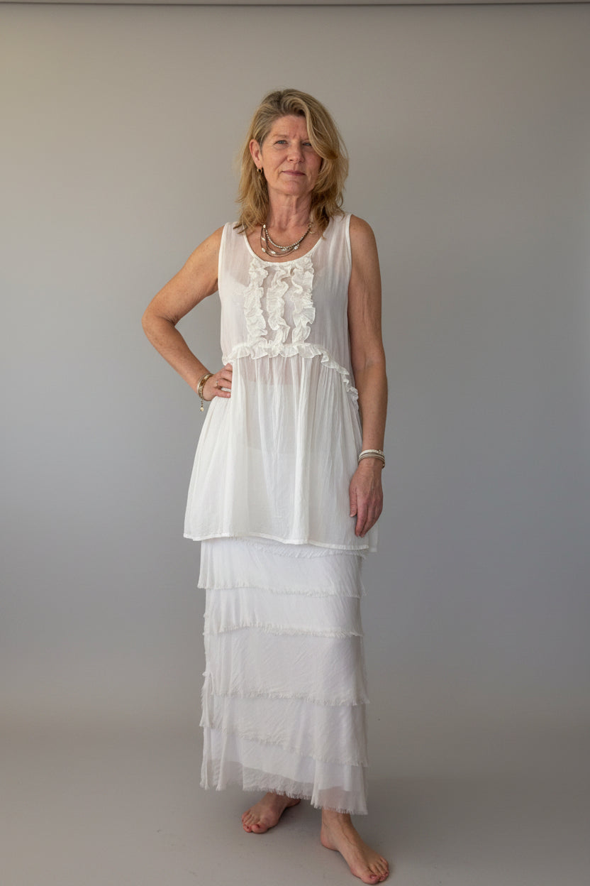 A woman stands barefoot in a room with a light grey wall, wearing a white sleeveless long cotton top with ruffles and a silk latte colored tiered skirt.  Cheeky Goddess.  Australia.
