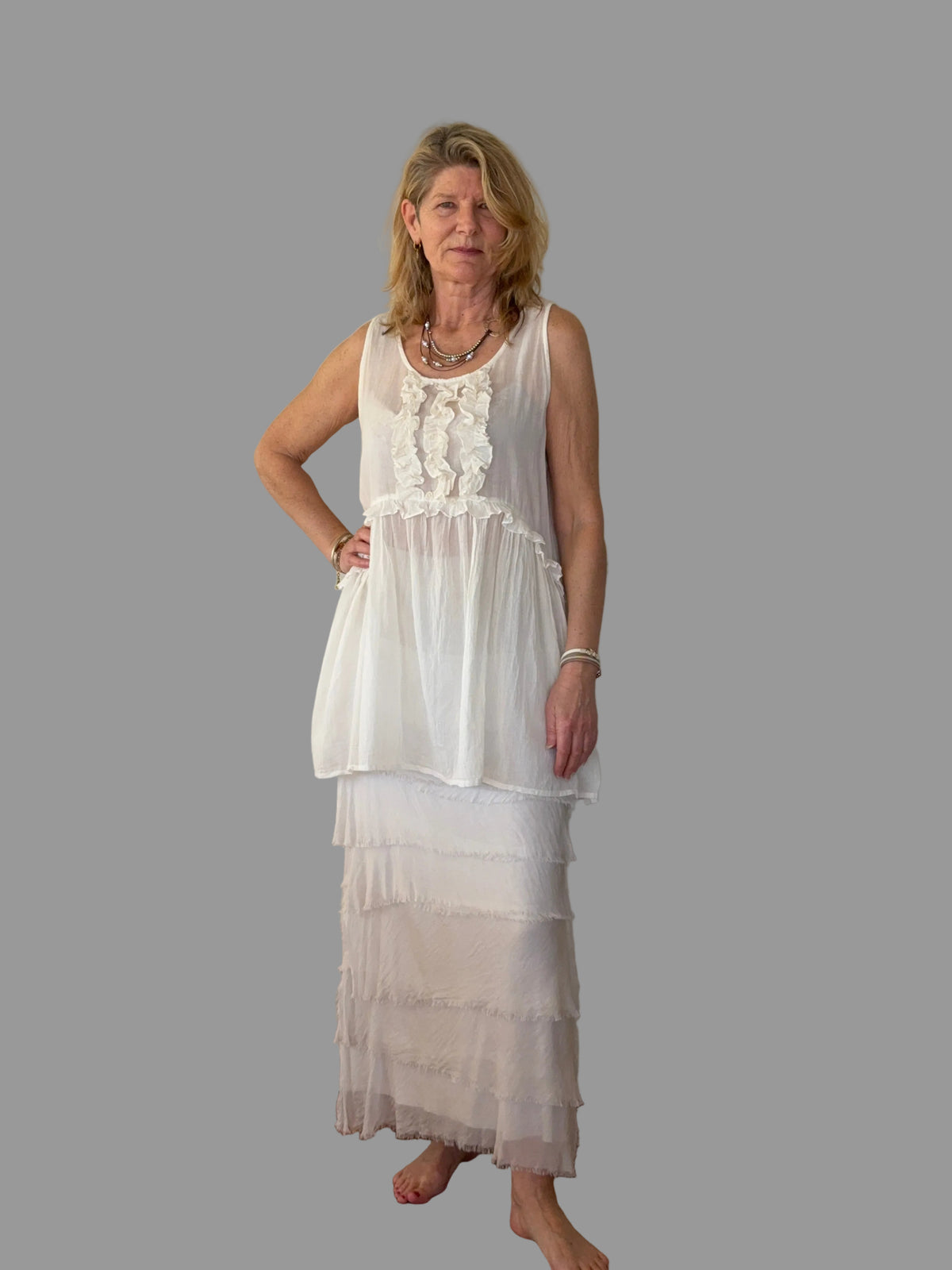 A woman stands barefoot in a room with a light beige wall and wooden floor, wearing a white sleeveless maxi dress with a ruffled tiered skirt.