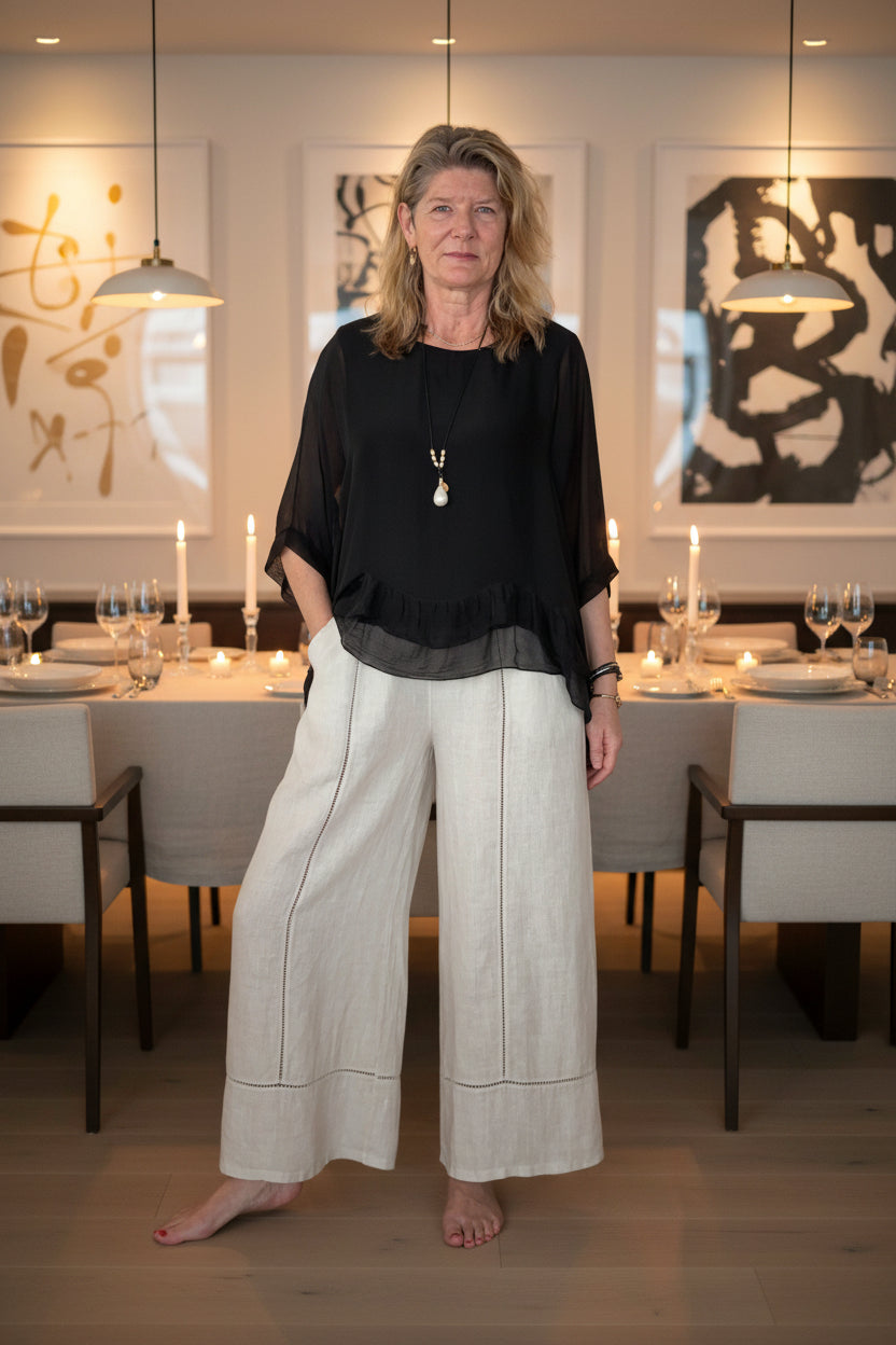 A woman stands barefoot on a wooden floor against a plain white wall, wearing a loose black short-sleeve top with a ruffled hem and wide-leg off-white trousers.  Cheeky Goddess.