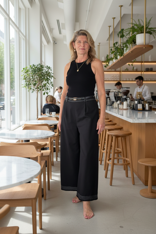 A woman stands barefoot in a bright, modern cafe or restaurant, wearing a sleeveless black top and black wide-leg cropped pants with a thin metallic belt.