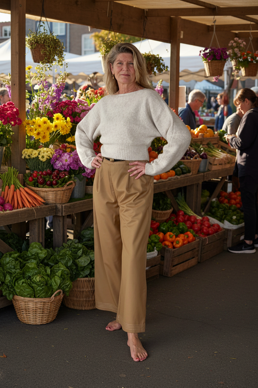 A woman stands barefoot at an outdoor farmers market wearing a cream-colored knit sweater and tan wide-leg trousers, posing with her hands on her hips in front of colorful flower and vegetable stalls.