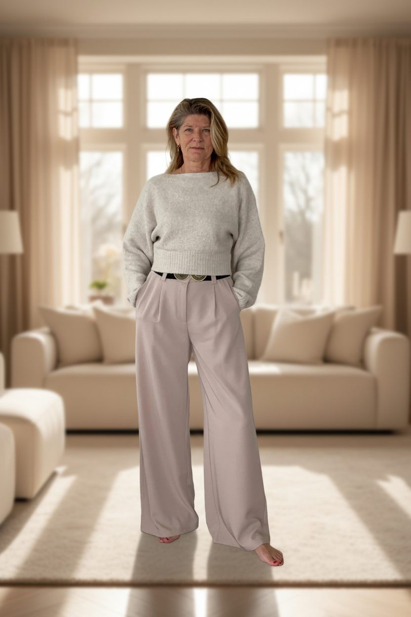 Woman standing in a bright living room wearing a light gray sweater and pants.