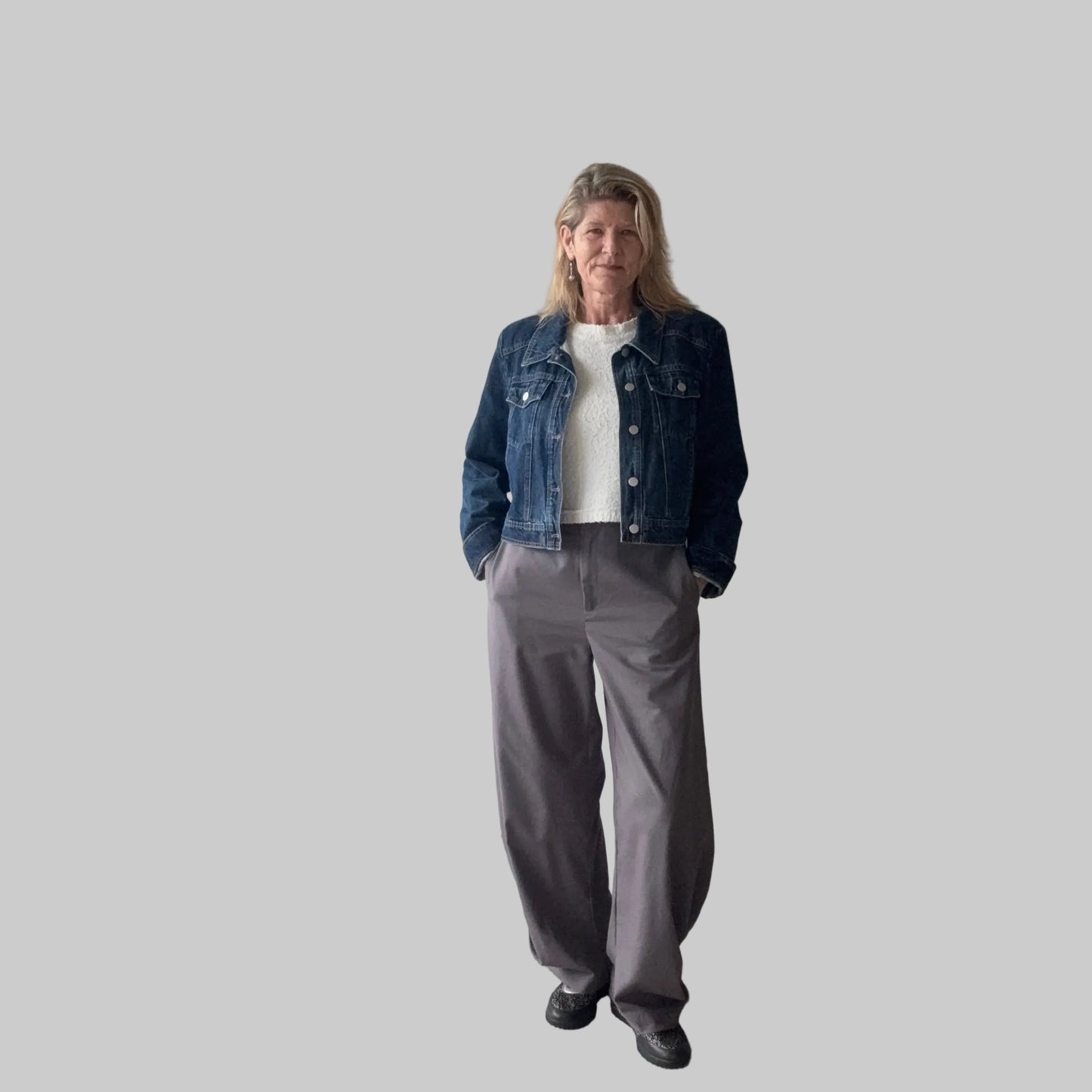 A woman stands indoors wearing a dark blue denim jacket over a white top, paired with loose gray wide-leg pants and black shoes. She is positioned in front of a plain beige wall next to a large window. Cheeky Goddess.