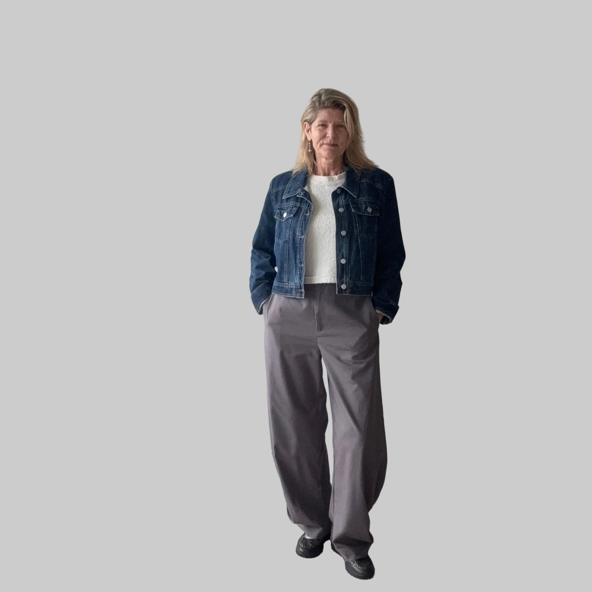 A woman stands indoors wearing a dark blue denim jacket over a white top, paired with loose gray wide-leg pants and black shoes. She is positioned in front of a plain beige wall next to a large window. Cheeky Goddess.