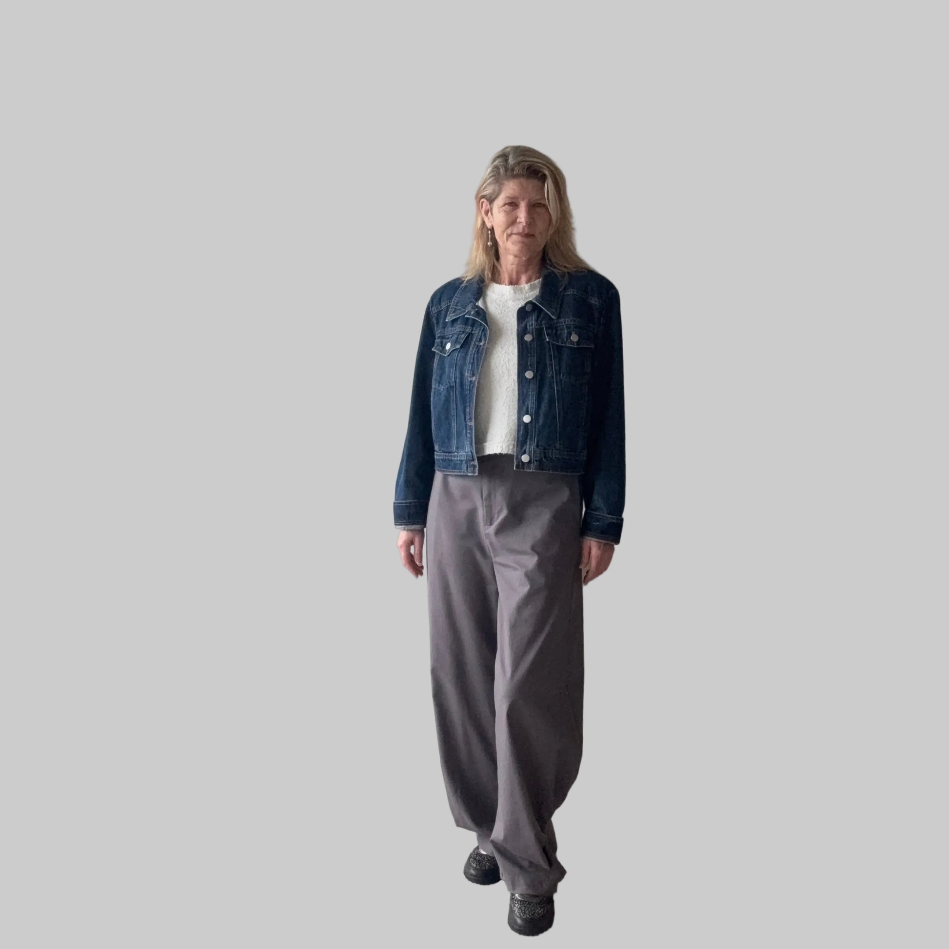A woman stands indoors wearing a dark blue denim jacket over a white top, paired with loose gray wide-leg pants and black shoes. She is positioned in front of a plain beige wall next to a large window. Cheeky Goddess