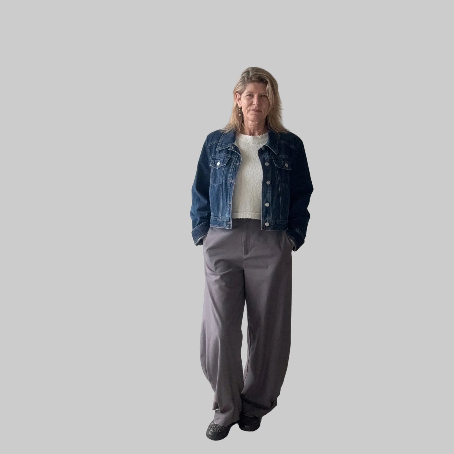 A woman stands indoors wearing a dark blue denim jacket over a white top, paired with loose gray wide-leg pants and black shoes. She is positioned in front of a plain beige wall next to a large window. Cheeky Goddess