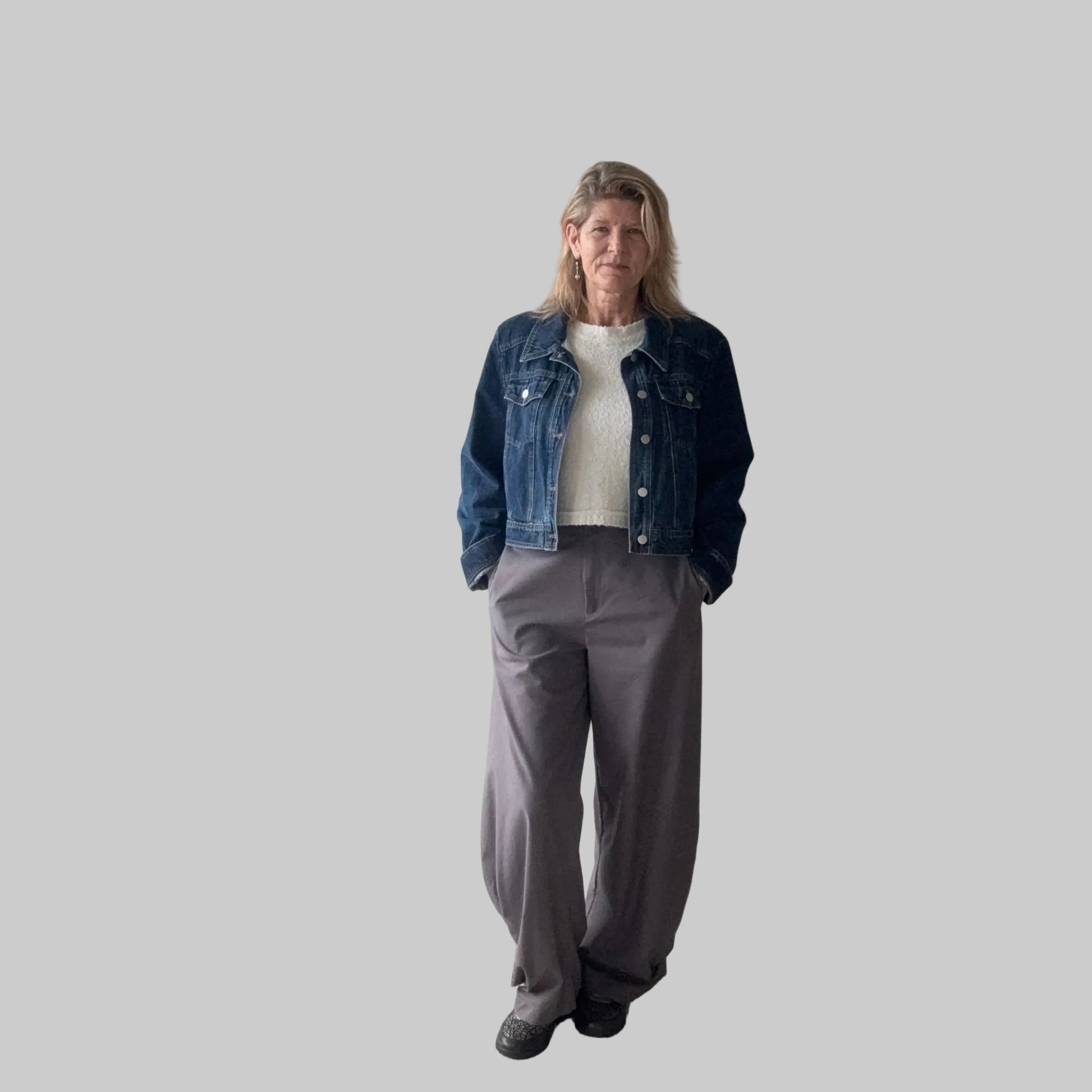 A woman stands indoors wearing a dark blue denim jacket over a white top, paired with loose gray wide-leg pants and black shoes. She is positioned in front of a plain beige wall next to a large window. Cheeky Goddess