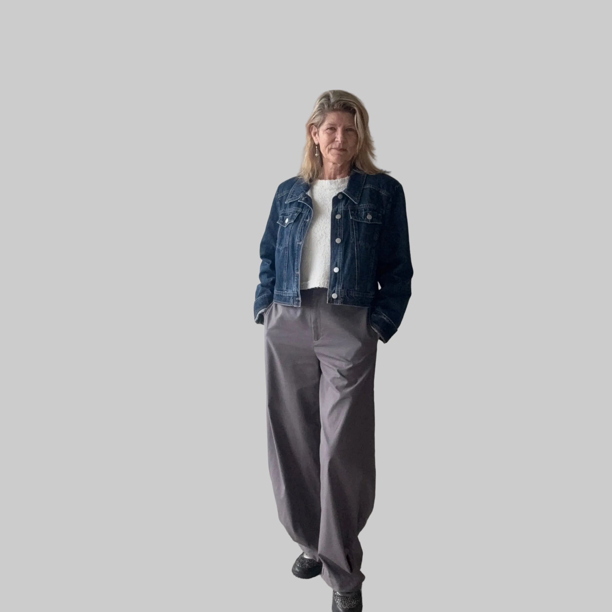 A woman stands indoors wearing a dark blue denim jacket over a white top, paired with loose gray wide-leg pants and black shoes. She is positioned in front of a plain beige wall next to a large window. Cheeky Goddess