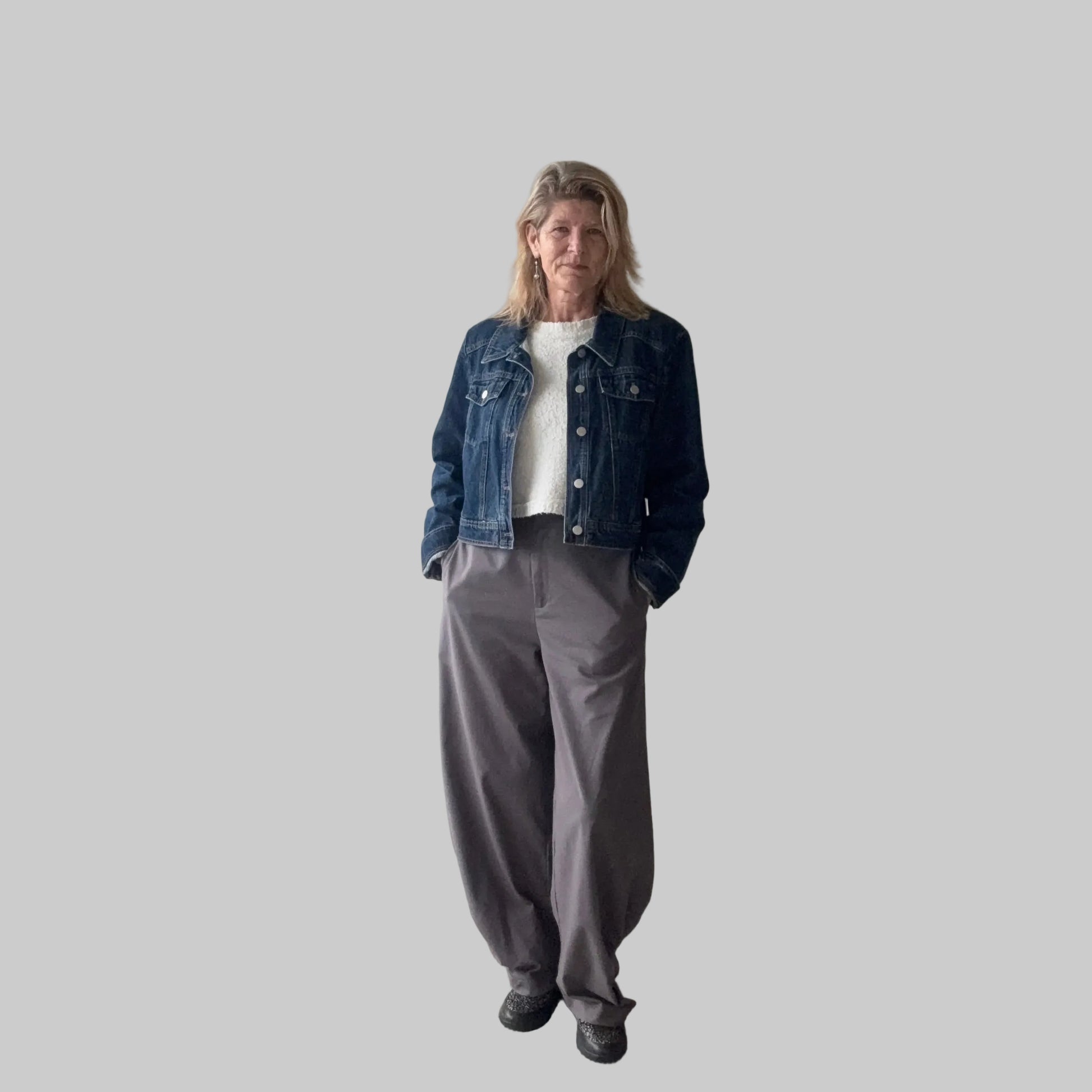 A woman stands indoors wearing a dark blue denim jacket over a white top, paired with loose gray wide-leg pants and black shoes. She is positioned in front of a plain beige wall next to a large window. Cheeky Goddess