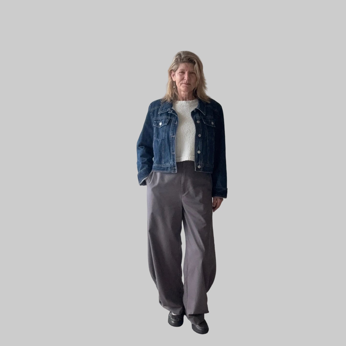 A woman stands indoors wearing a dark blue denim jacket over a white top, paired with loose gray wide-leg pants and black shoes. She is positioned in front of a plain beige wall next to a large window. Cheeky Goddess