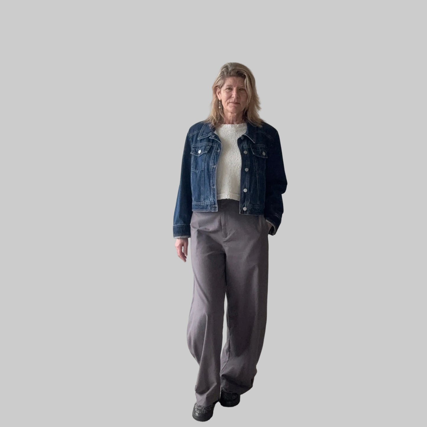 A woman stands indoors wearing a dark blue denim jacket over a white top, paired with loose gray wide-leg pants and black shoes. She is positioned in front of a plain beige wall next to a large window. Cheeky Goddess