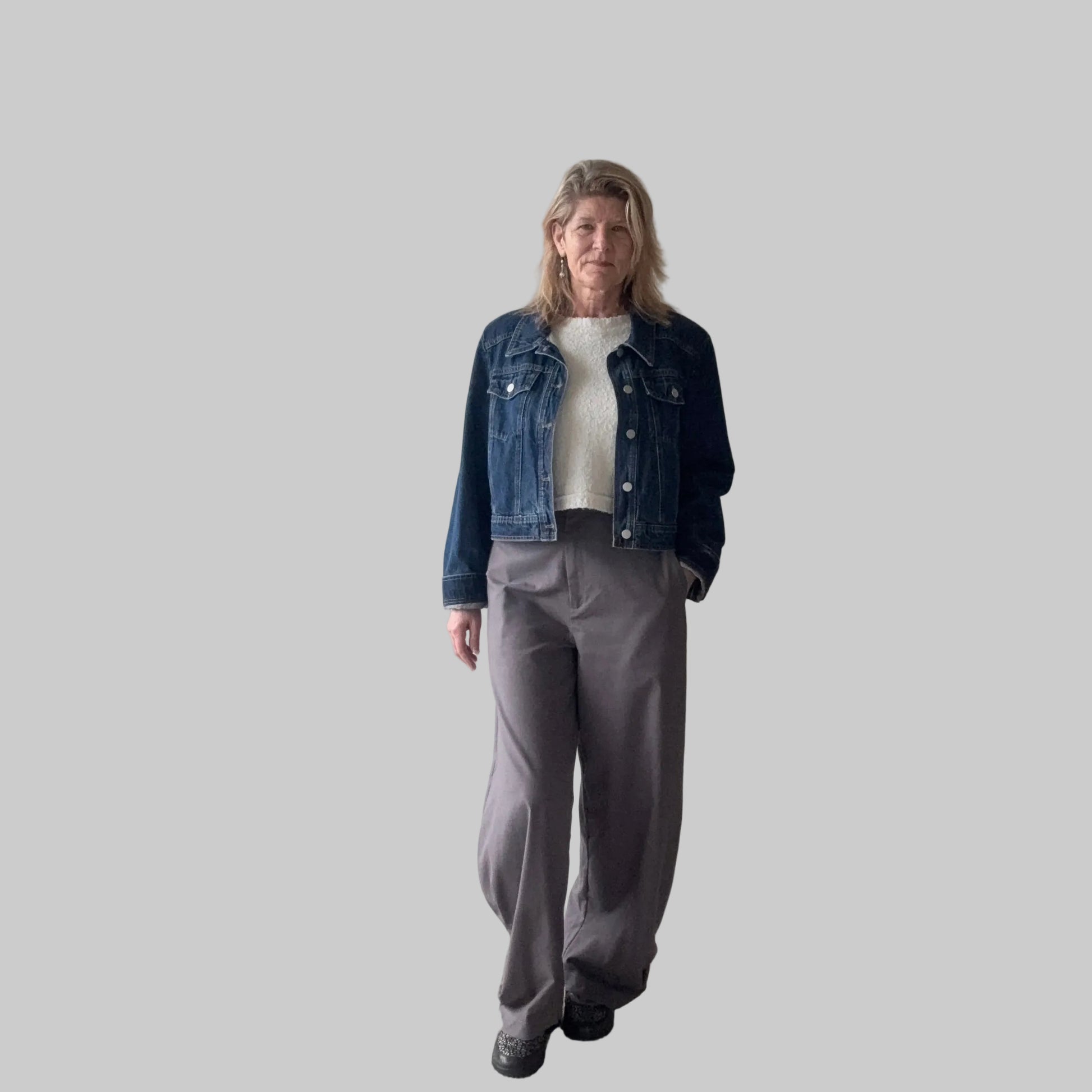A woman stands indoors wearing a dark blue denim jacket over a white top, paired with loose gray wide-leg pants and black shoes. She is positioned in front of a plain beige wall next to a large window. Cheeky Goddess