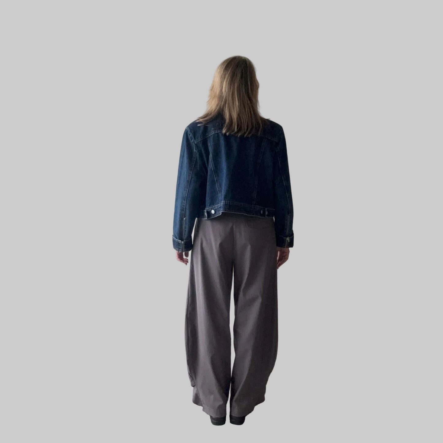 A woman standing with their back to the camera, wearing a dark blue denim jacket and loose, wide-leg gray pants with a plain light grey background. Cheeky Goddess.