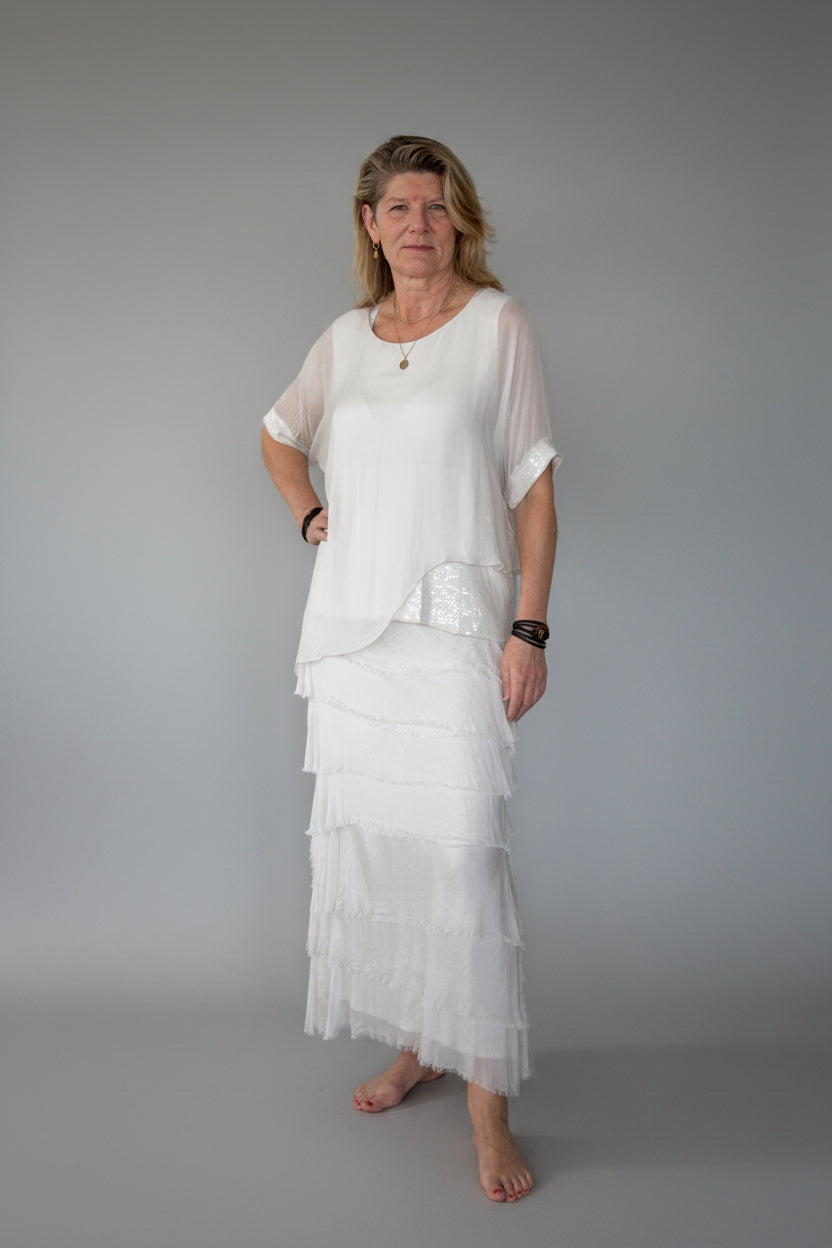 A woman stands barefoot against a plain off-white wall on a wooden floor, wearing a white layered silk tunic top with short sleeves and a long tiered maxi silk skirt.  Cheeky Goddess.  Australia