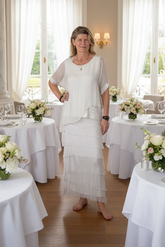 A woman stands barefoot in a bright, elegant dining room wearing a white tiered tunic top and matching long skirt. The room features white tablecloths, floral arrangements, and large windows with sheer curtains.  Silk top and skirt.  Cheeky Goddess.  Australia.